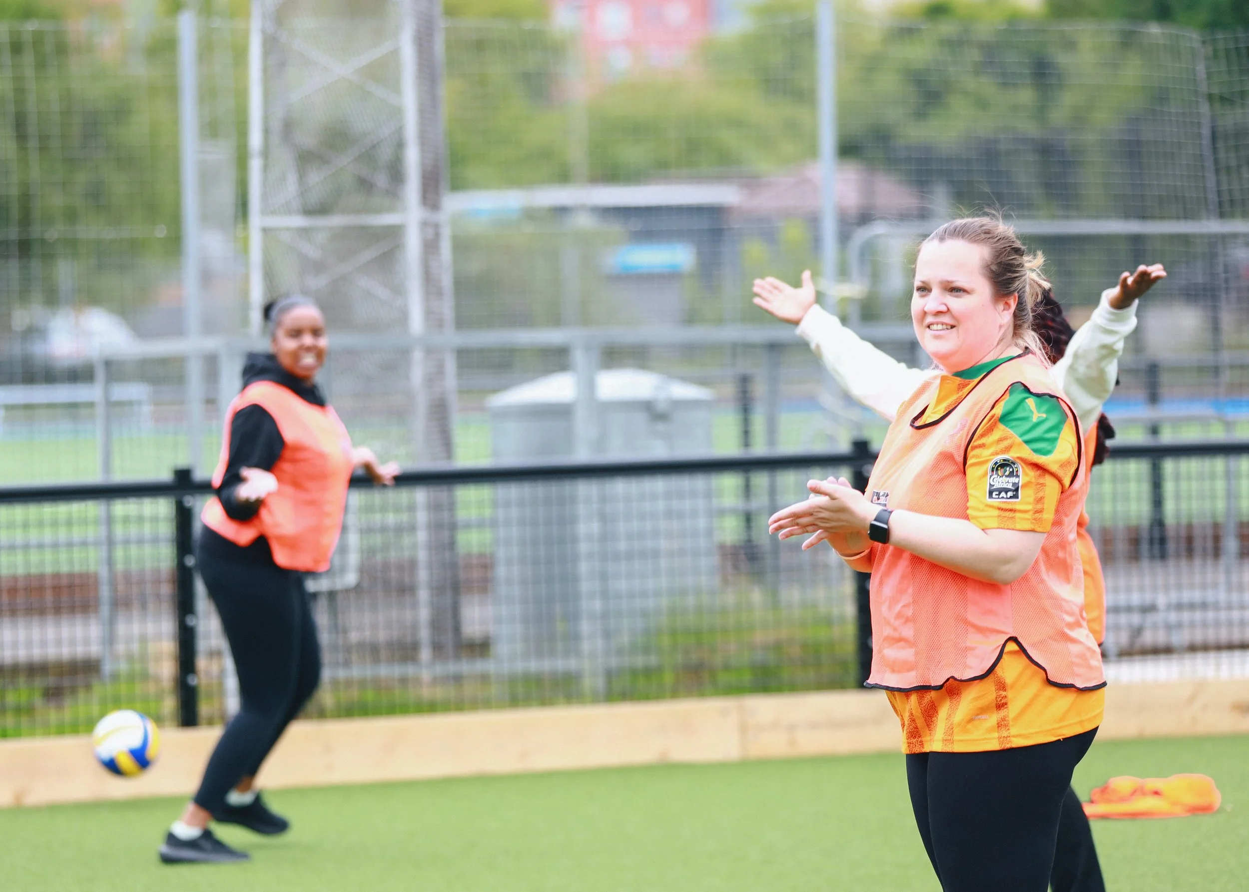 Women playing volleyball on an outdoor court, wearing orange vests and engaging in a game.