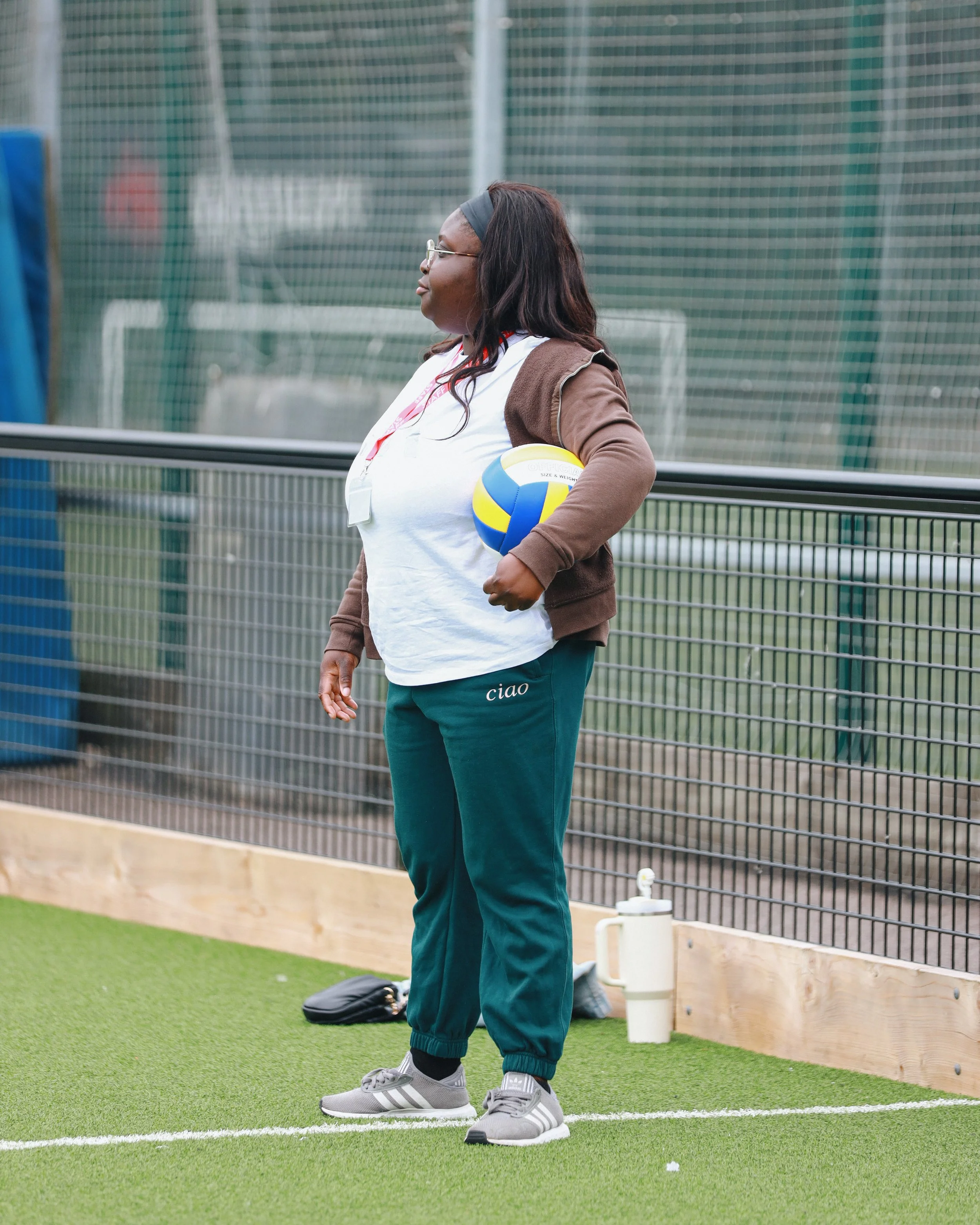 A person standing on a sports field, holding a volleyball in their right hand, wearing athletic clothes including a white shirt, black pants with 'ciao' written on the left leg, and gray sports shoes, with sunglasses and a headband.