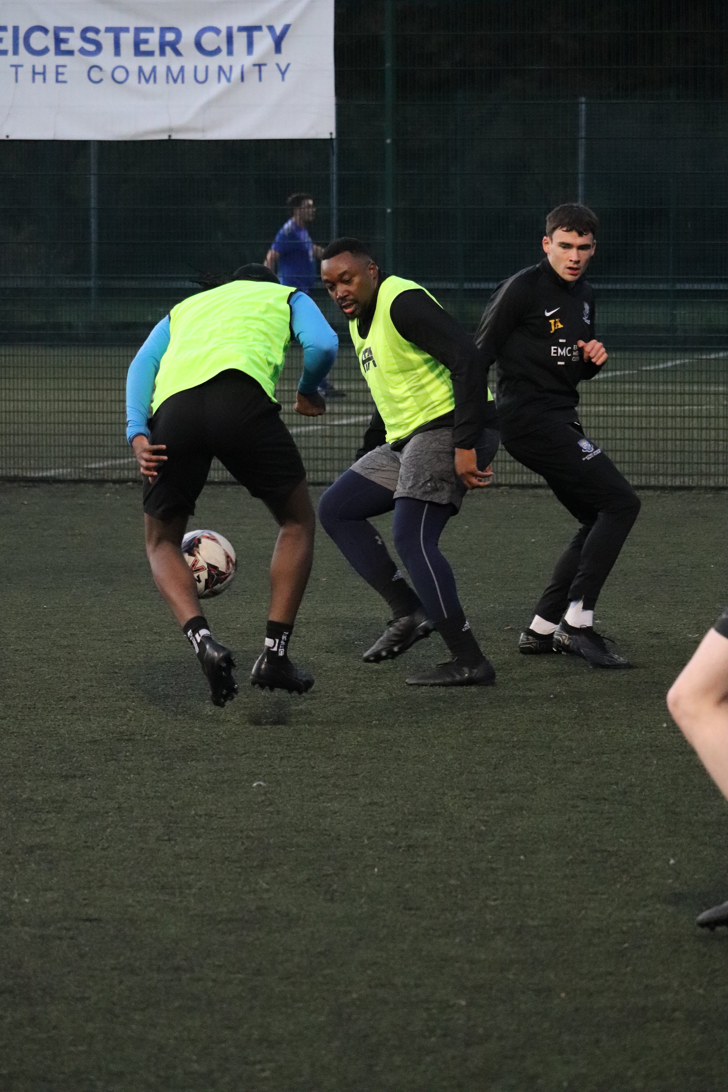 Three soccer players are engaged in a practice on a field, with one player kicking the ball while the other two observe or prepare to intercept, surrounded by a fence and a banner in the background.