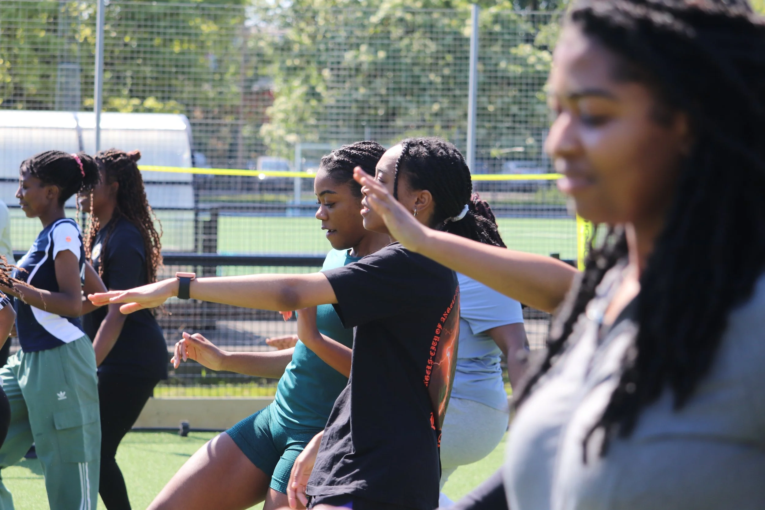 Group of women practicing yoga outdoors on a sports field with a fence in the background.