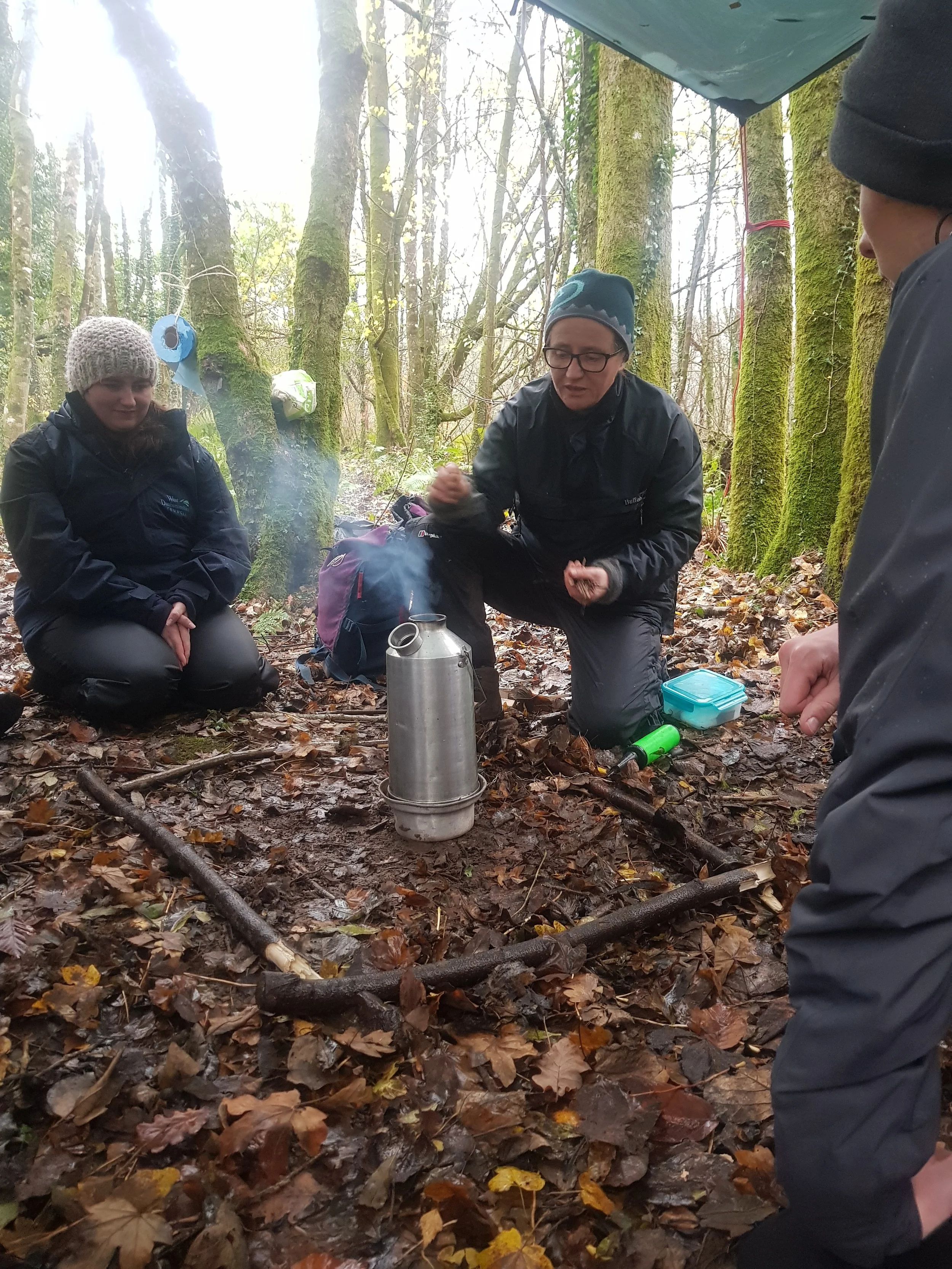 A group of people in outdoor clothing sitting and kneeling in a forest clearing with wet leaves on the ground, gathered around a small fire with a metal kettle, with some camping gear visible.