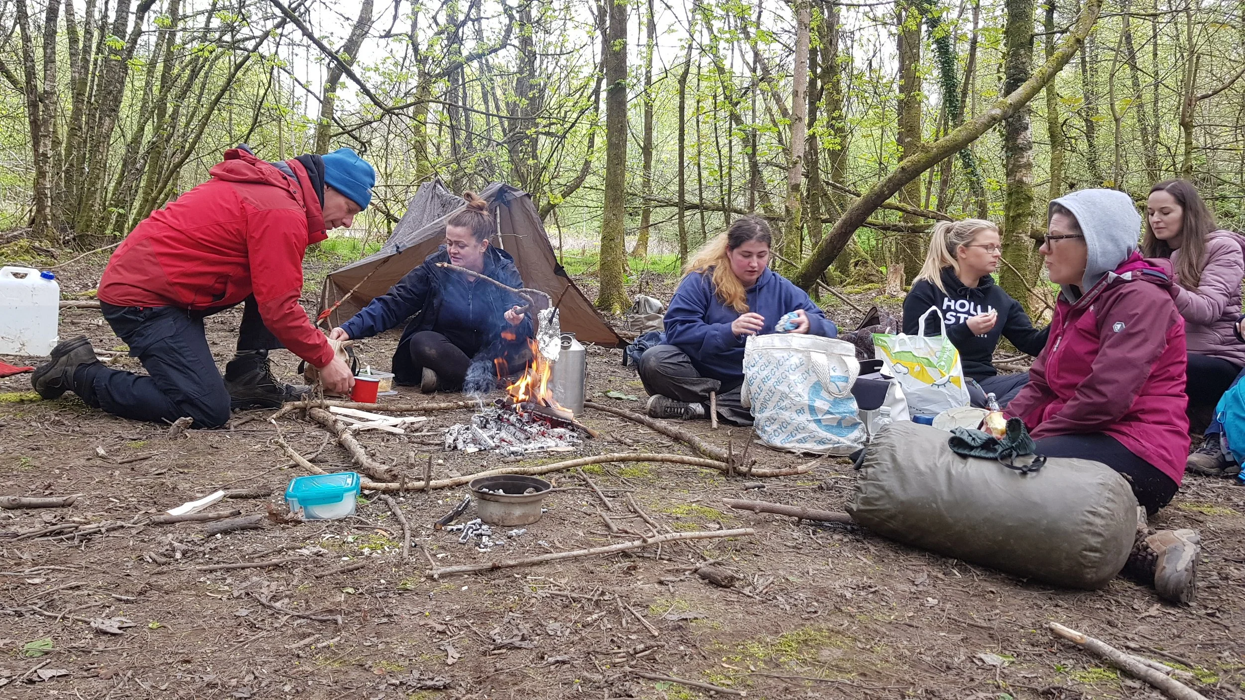 Group of people sitting and kneeling around a campfire in a forest, with camping gear and trees in the background.