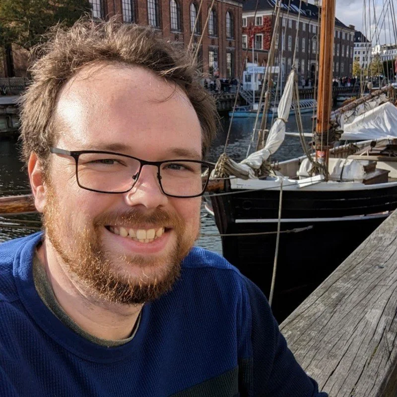 A man with glasses and a beard smiling at the camera, with boats and brick buildings along a canal in the background.