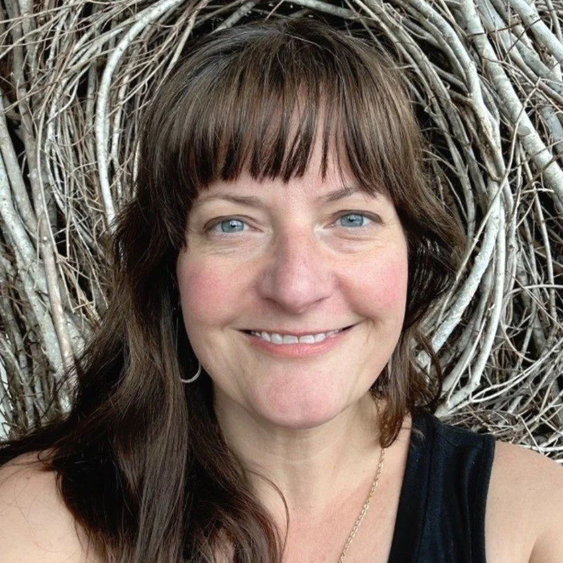 A woman with brown hair and blue eyes smiling in front of a large nest made of twigs.