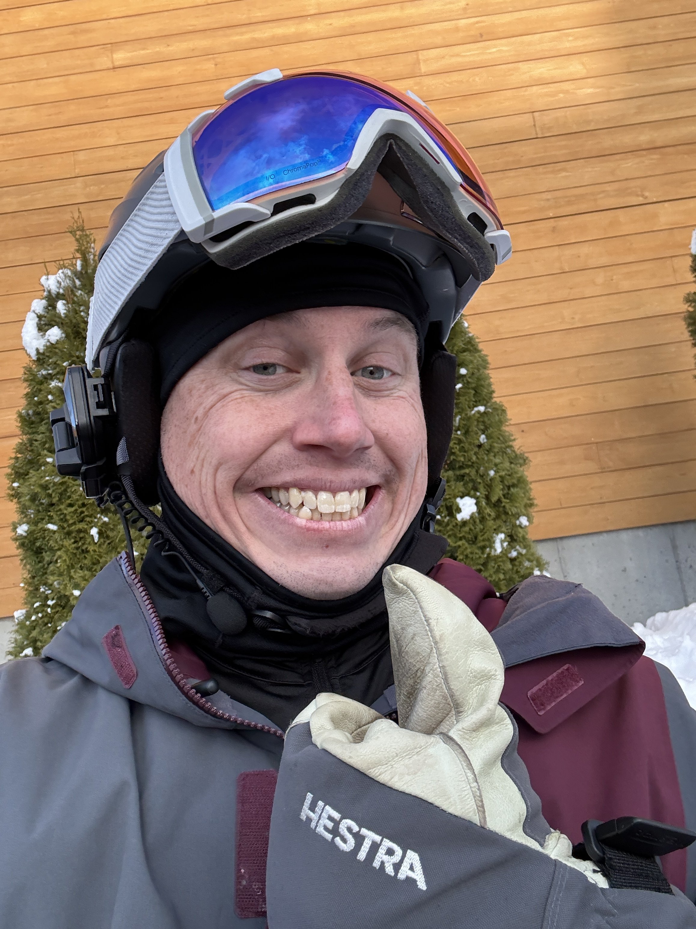 Portrait of a man in a snow gear smiling outdoors with green foliage in the background.