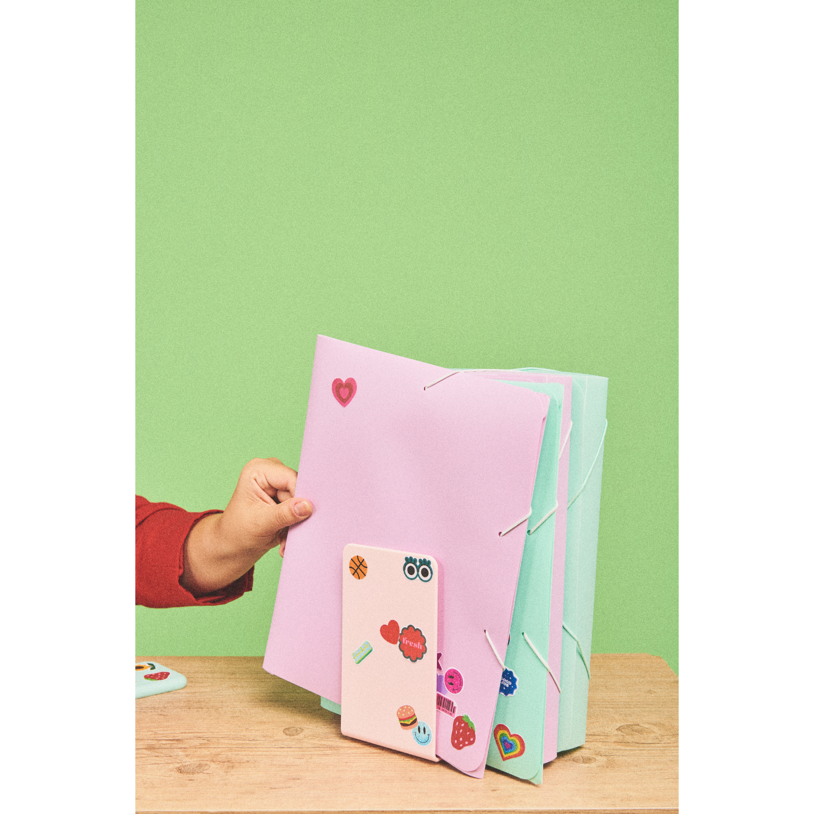 A hand holding a pink folder with stickers, next to green and pink folders tied with elastic bands on a wooden surface with a light green background.