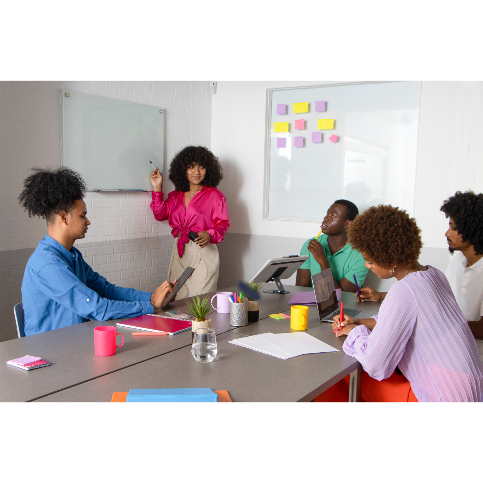 A diverse group of five people in a meeting room, with one woman standing and presenting at a whiteboard covered with colorful sticky notes, while others are seated around a table with laptops, notebooks, and writing materials.