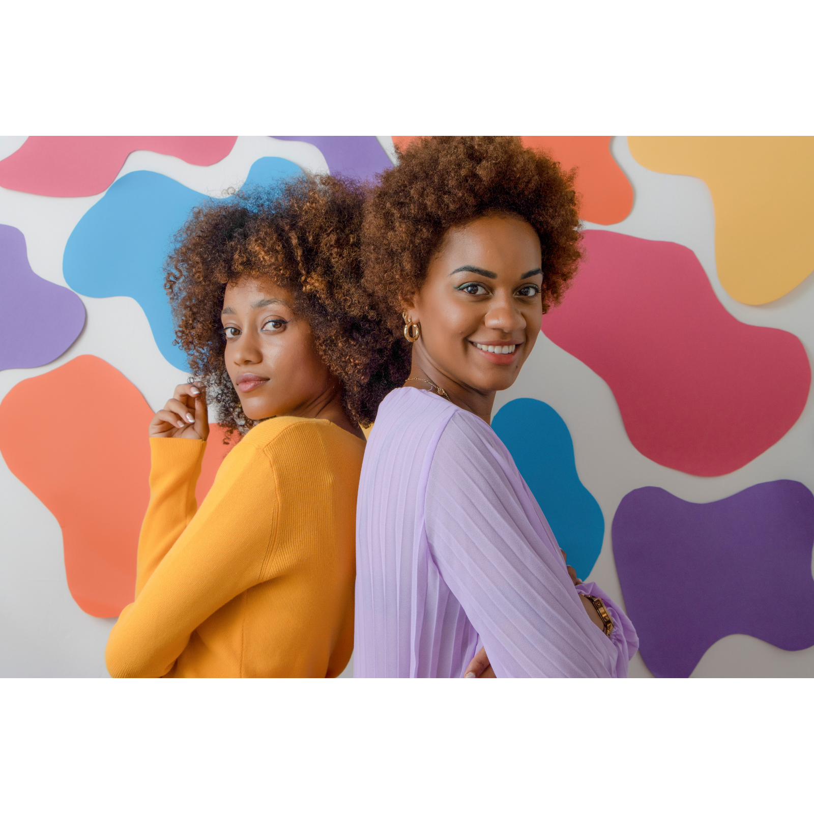 Two women with curly hair posing with their backs against each other, smiling, in front of a colorful abstract wall.