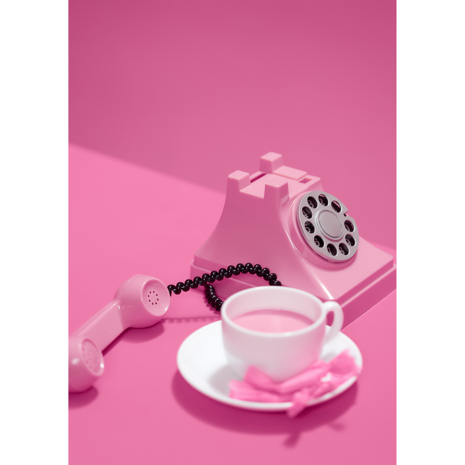 Pink rotary telephone with a black cord, white coffee cup on a saucer with a pink spoon and napkin, all set against a pink background.