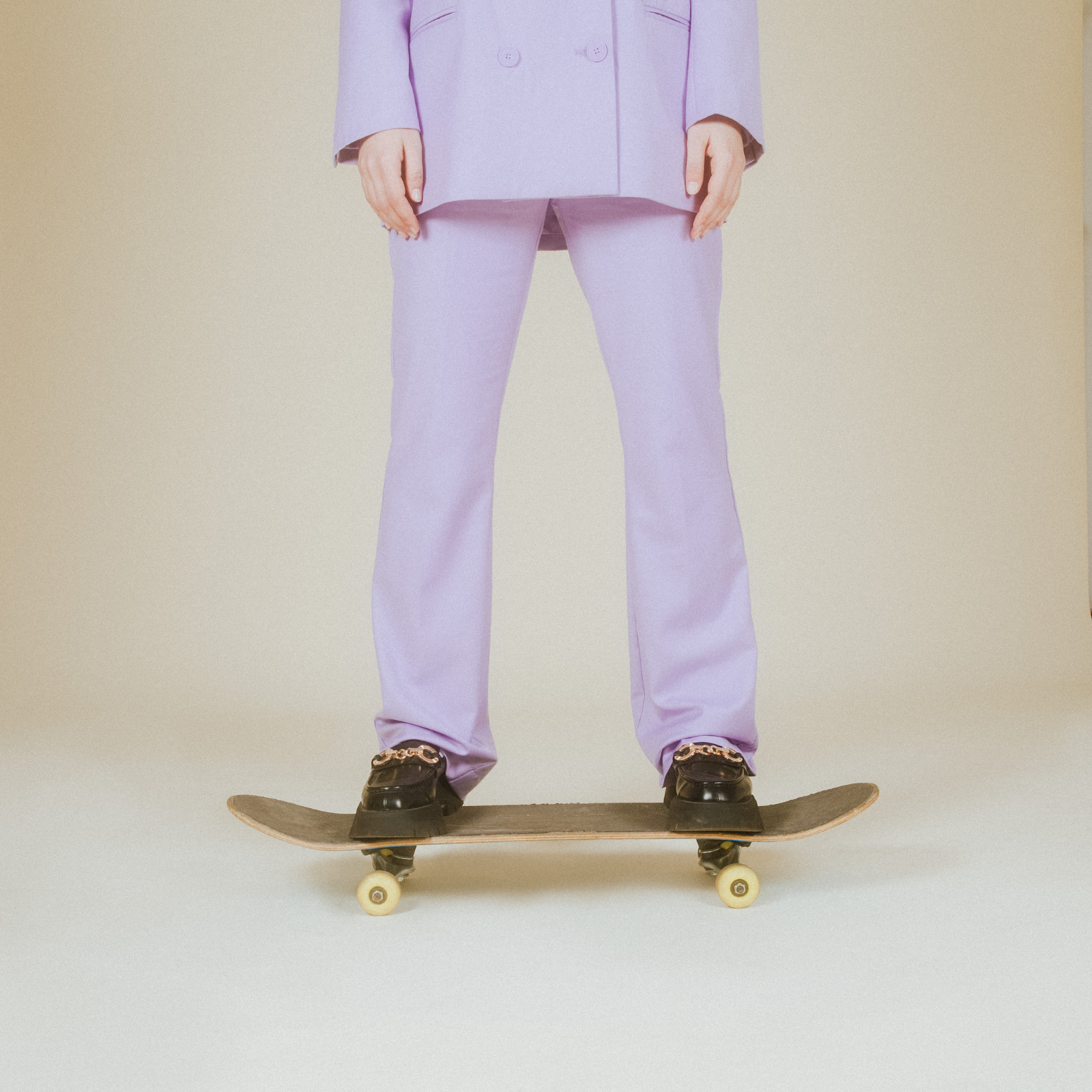 Person in purple pants and blazer standing on skateboard against beige background.