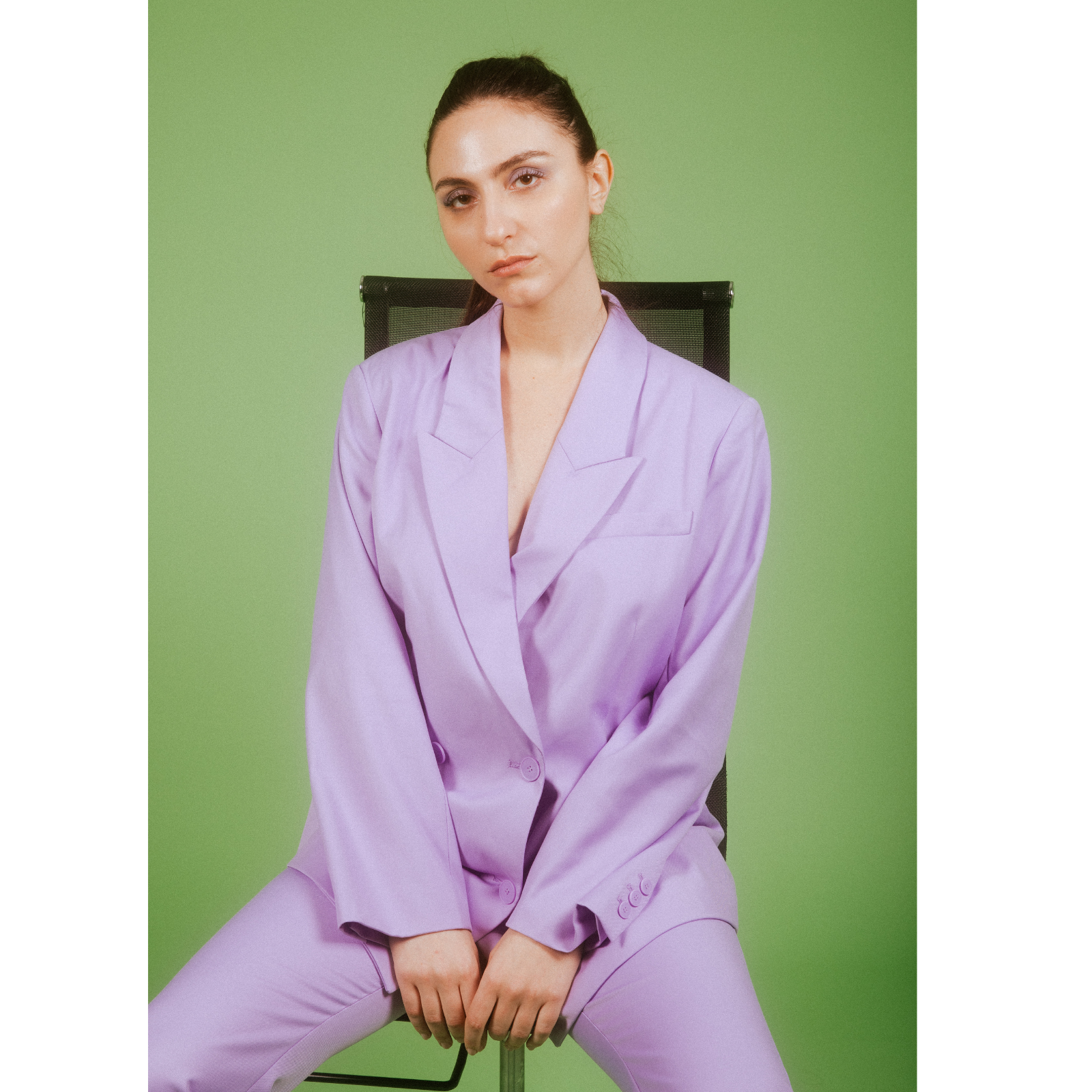 A woman in a lavender suit sitting on a black chair against a green background, looking directly at the camera with a neutral expression.