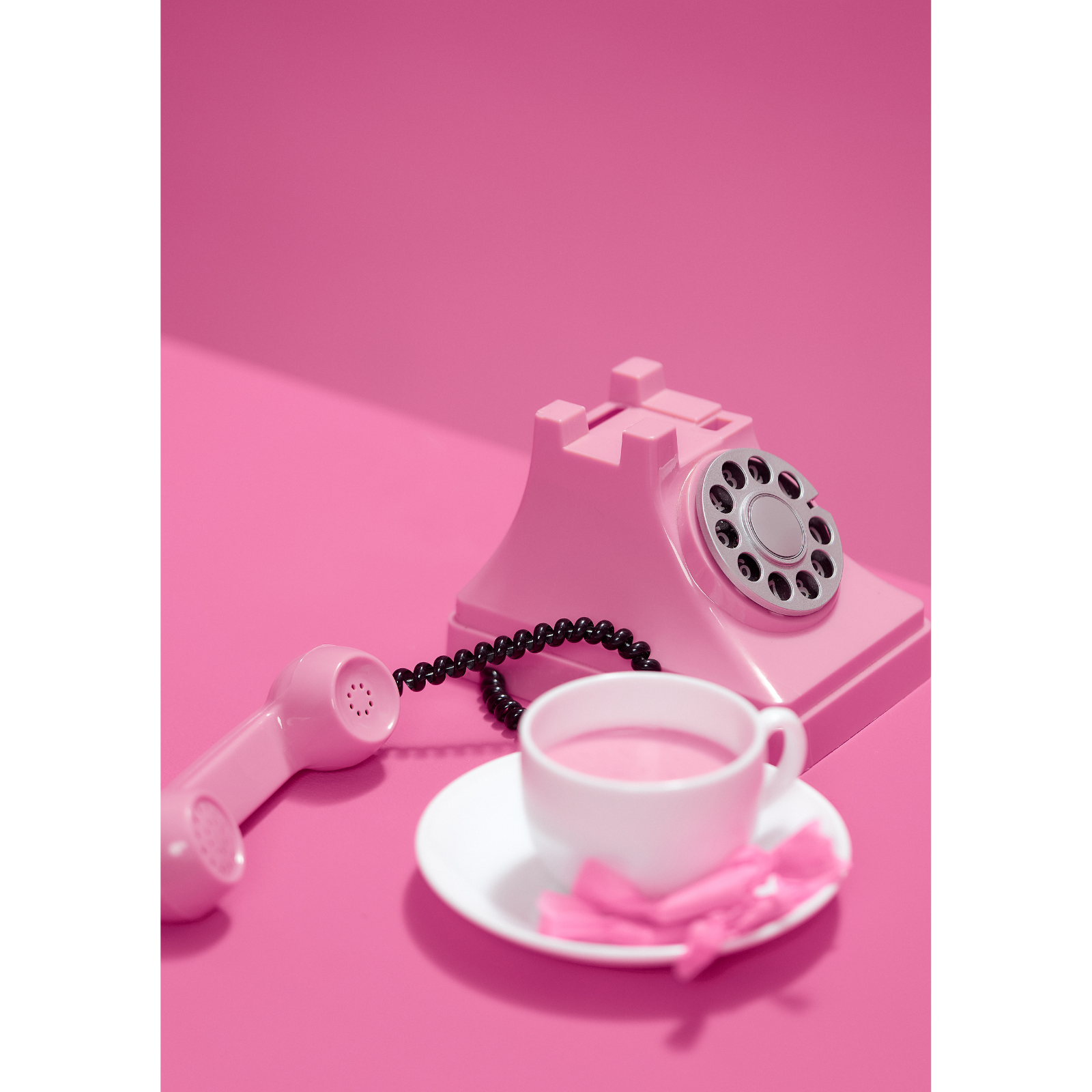 A pink rotary telephone, a white cup of tea on a saucer with a pink napkin, and a pink toy-like object on a pink surface with a pink background.