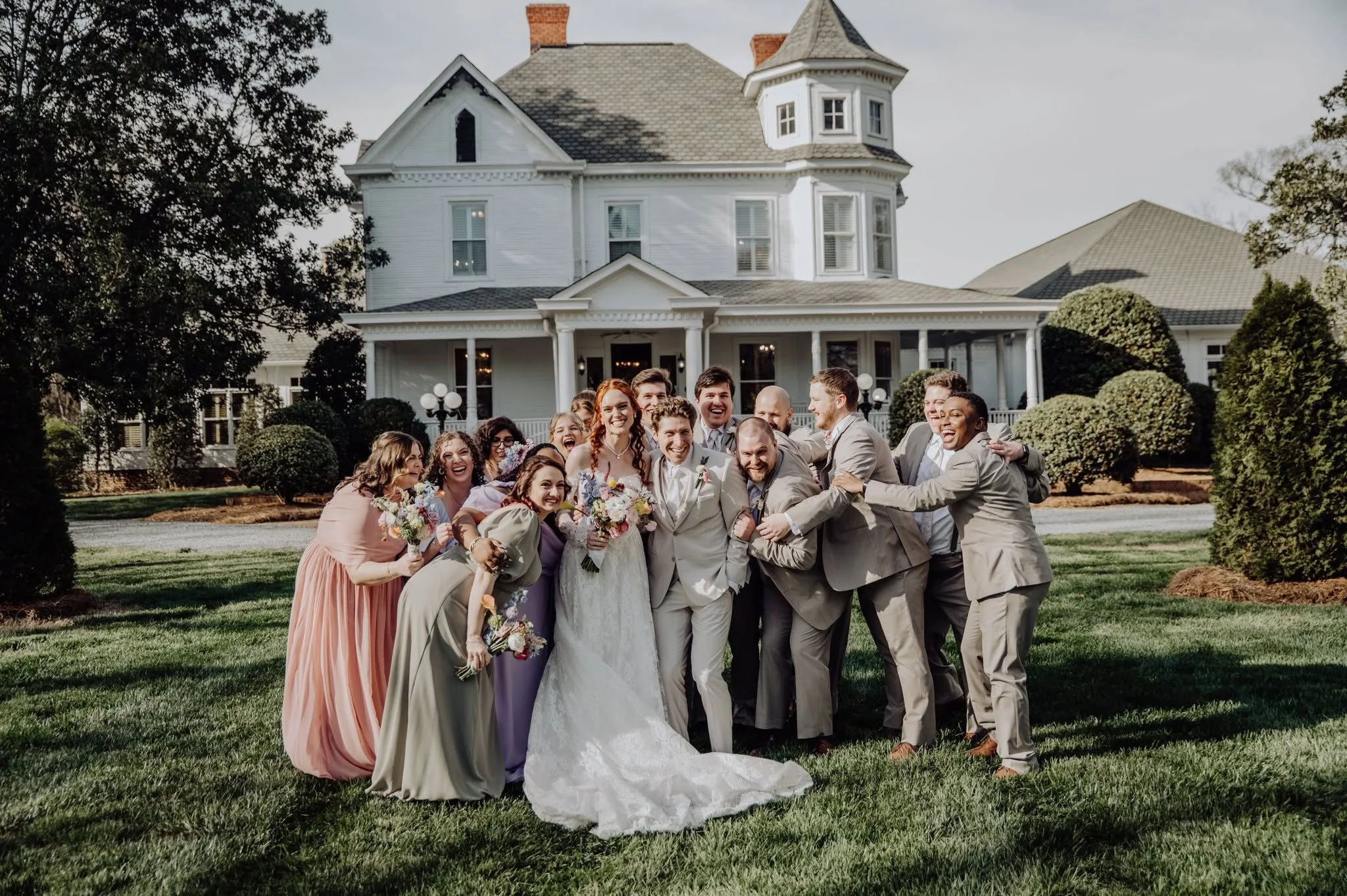 A large group of wedding guests gathered in front of a white Victorian-style house, smiling and celebrating with the bride and groom.