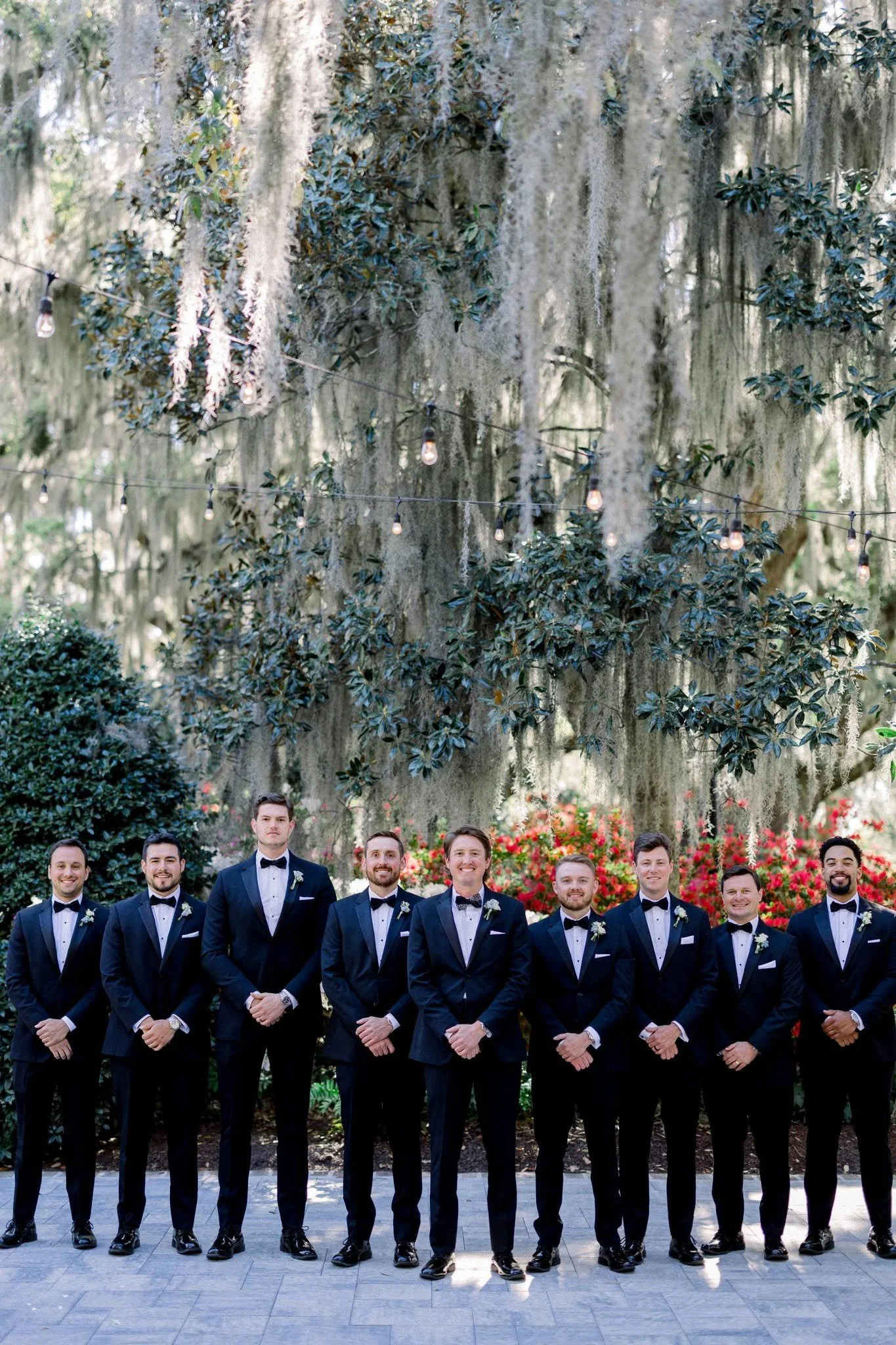 A group of eleven men dressed in black tuxedos and bow ties, standing outdoors in front of lush trees and vibrant red flowers, posing for a formal photo.