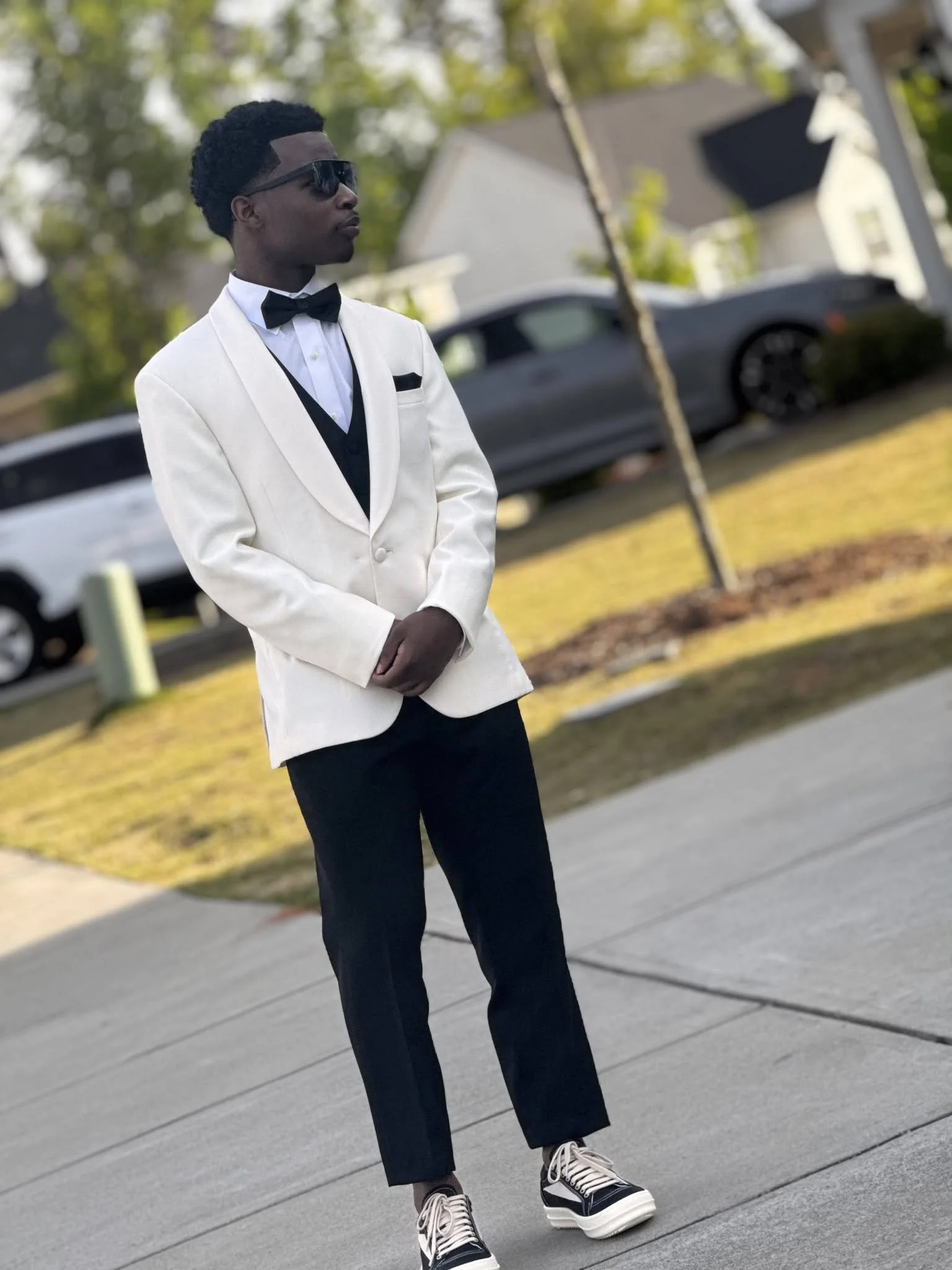 A young man in a white tuxedo jacket, black pants, and sneakers stands outdoors with trees and houses in the background. He is wearing sunglasses, a black bow tie, and has short curly hair.
