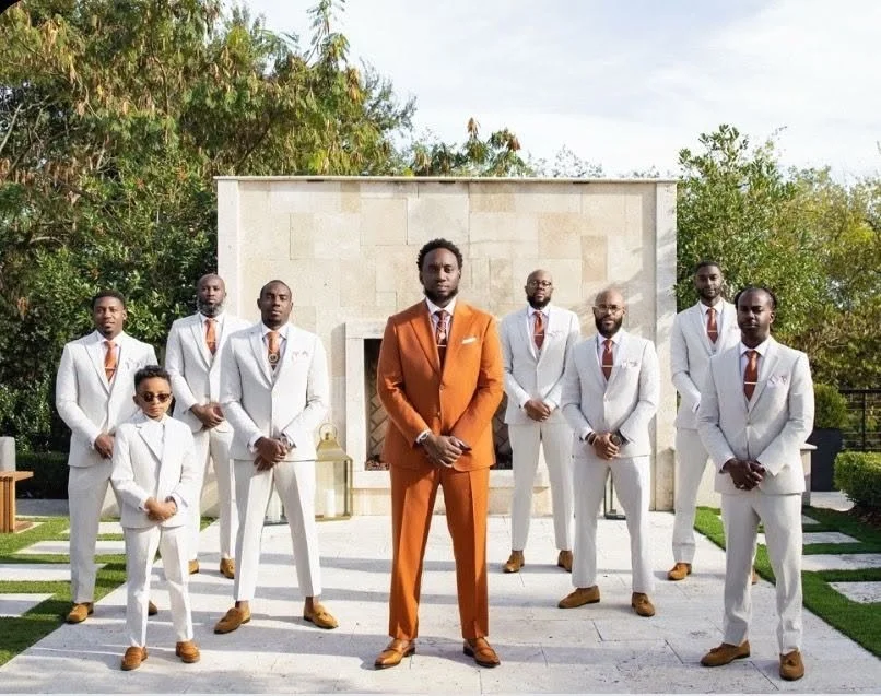 A group of nine men and one young boy dressed in matching white suits with orange ties and brown shoes, standing outdoors in front of a modern stone structure surrounded by greenery. The central man is wearing an orange suit.