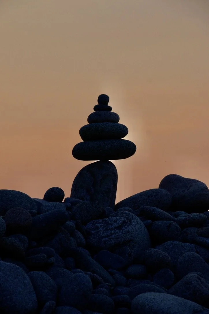 Stacked smooth black stones forming a balanced tower on a bed of rocks against a gradient sunset sky.