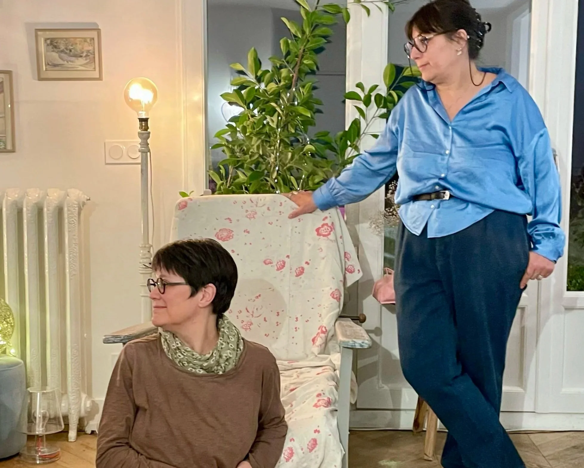 Deux femmes dans une pièce bien éclairée, l'une assise, portant des lunettes, et l'autre debout, touchant un fauteuil couvert d'une nappe blanche avec des motifs floraux, dans un intérieur avec des plantes, une lampe et des tableaux aux murs.