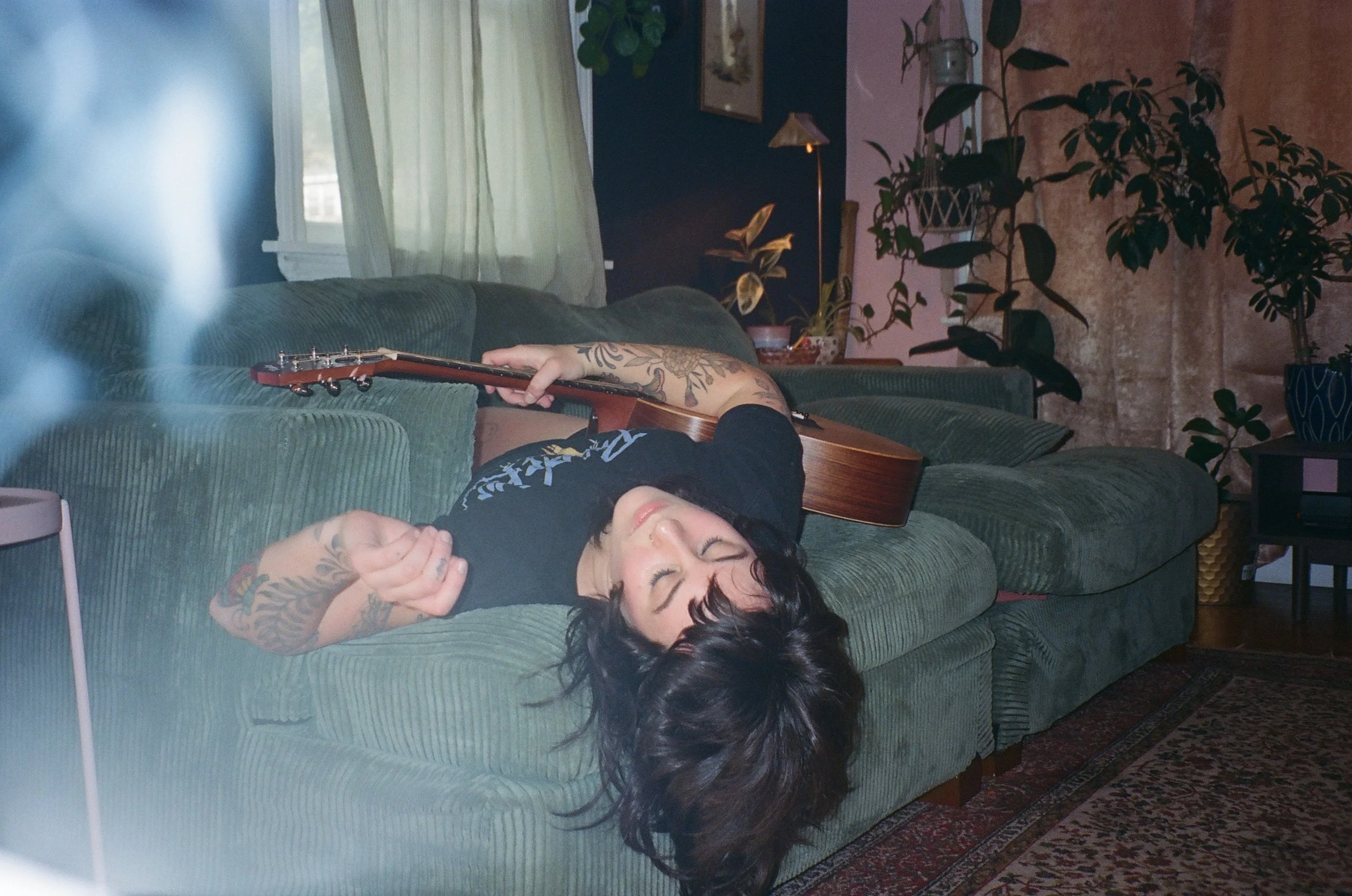 A woman with dark hair and tattoos is lying on a green couch, holding an acoustic guitar, with her eyes closed and a peaceful expression.