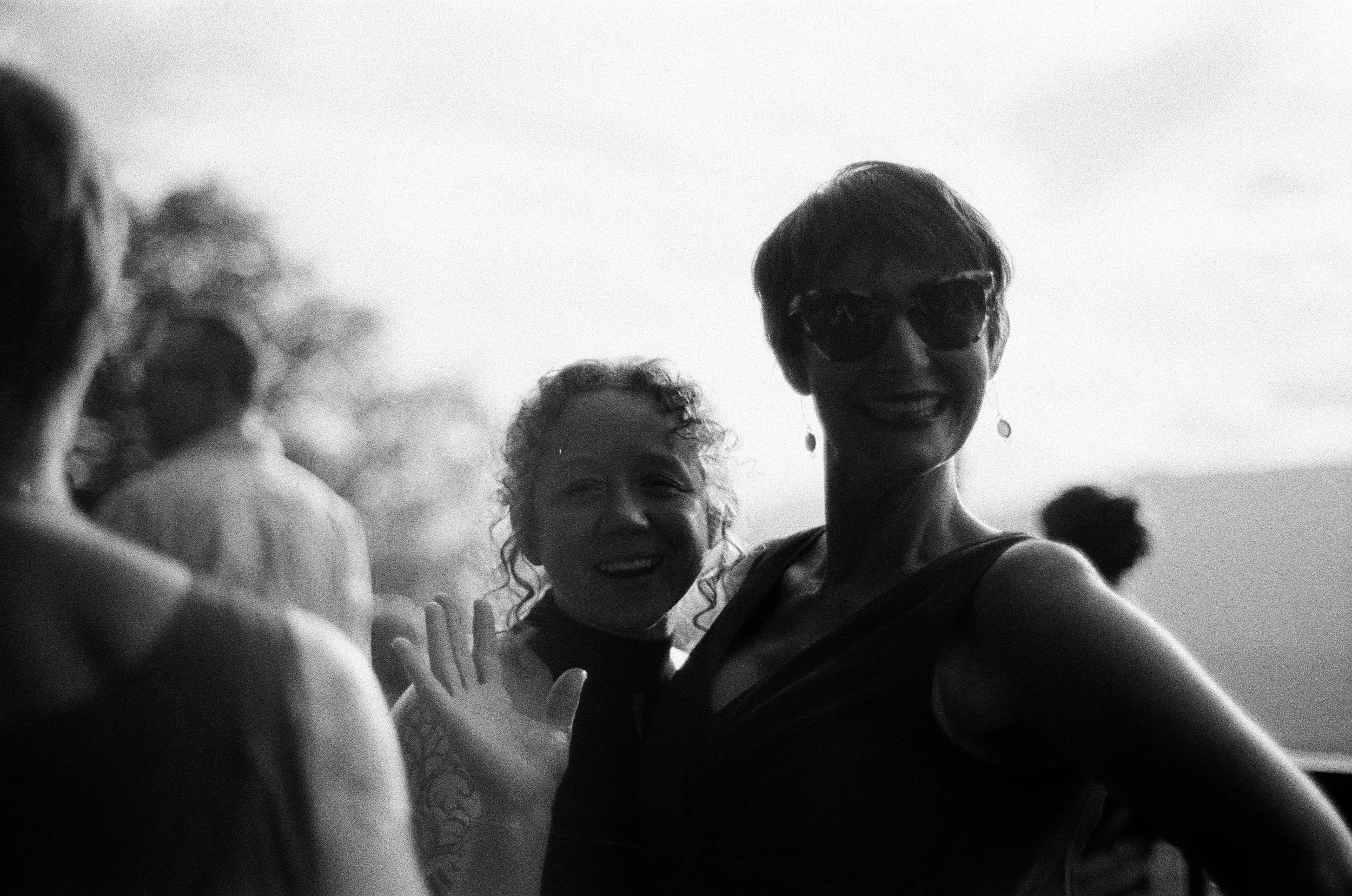Black and white photo of smiling women, one with short hair and sunglasses, waving at the camera, with others in the background outdoors.