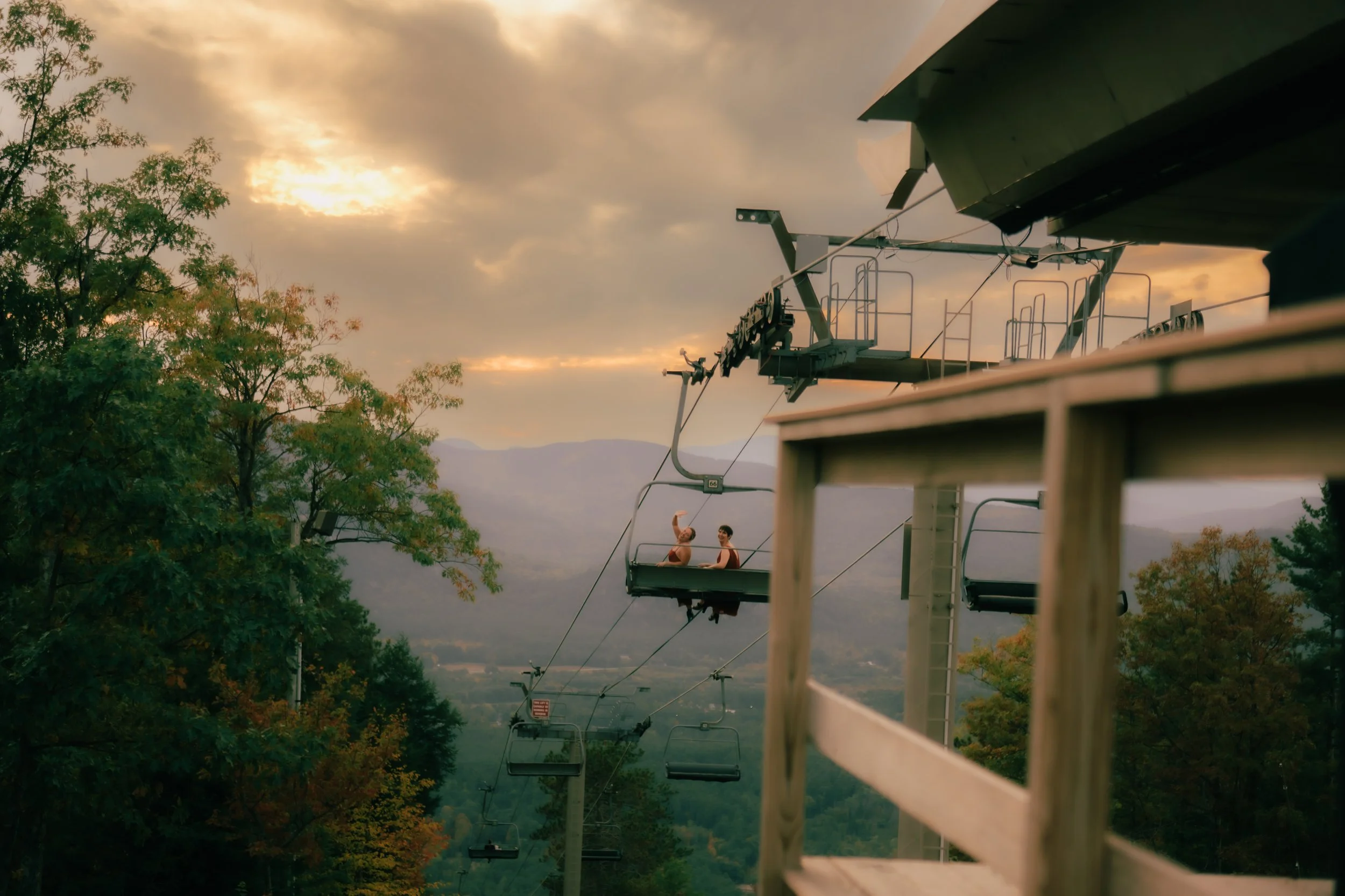 Two people sitting on a ski lift with trees and mountains in the background during sunset.