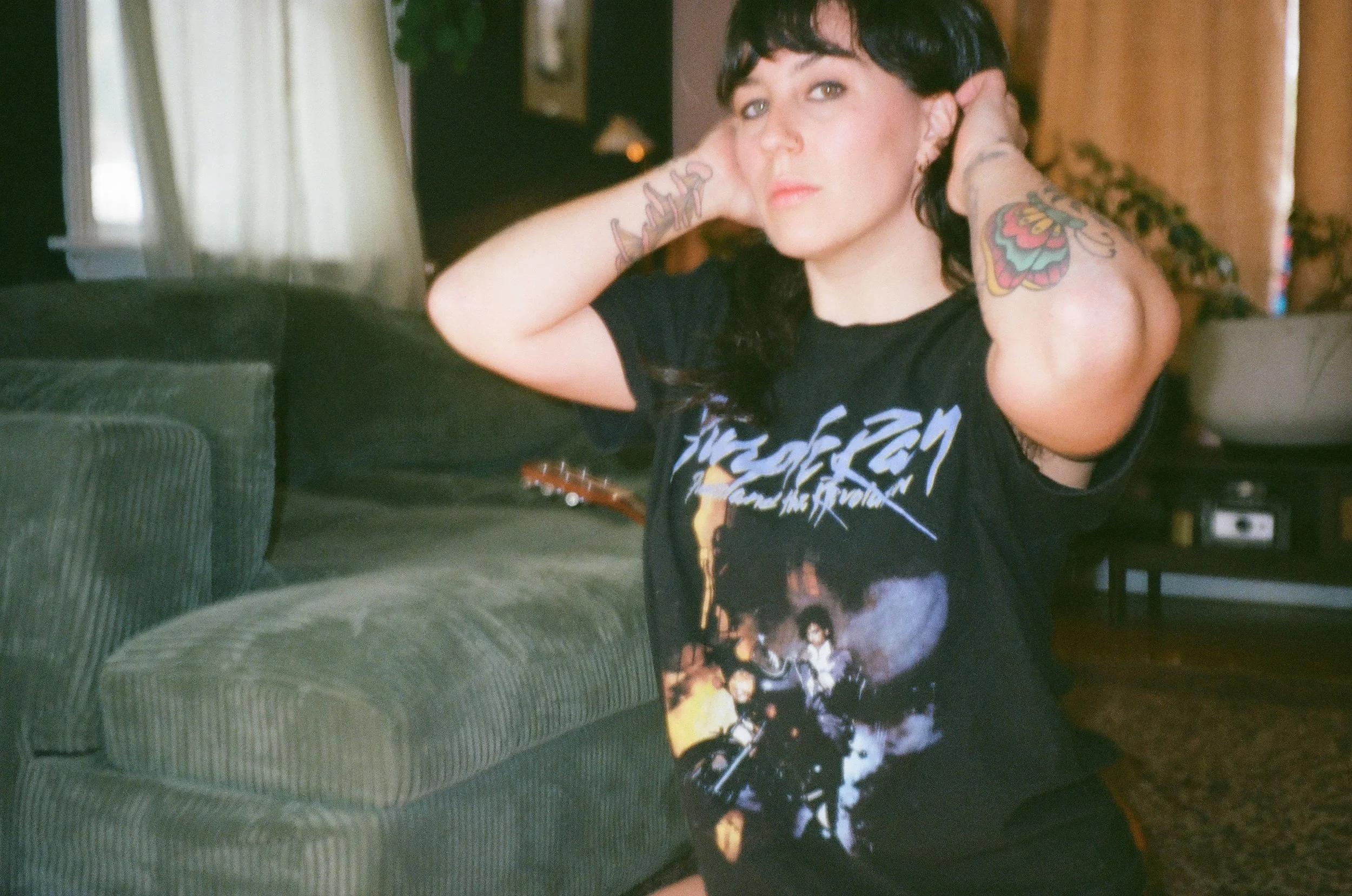 A woman with black hair and tattoos on her arms, wearing a black graphic T-shirt, is sitting on the floor in a room with a green sofa and brown curtains, adjusting her hair.