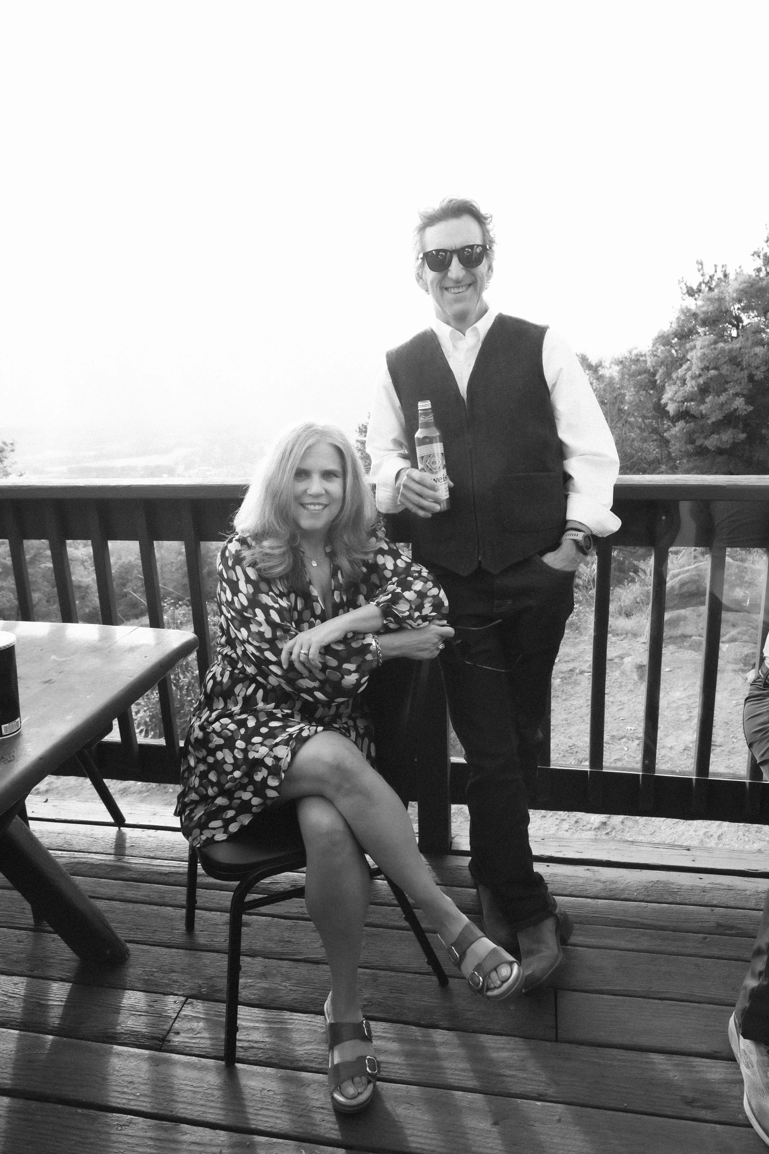 A man and woman enjoying time outdoors on a wooden deck, with the woman sitting on a chair and the man standing beside her holding a bottle, both smiling.