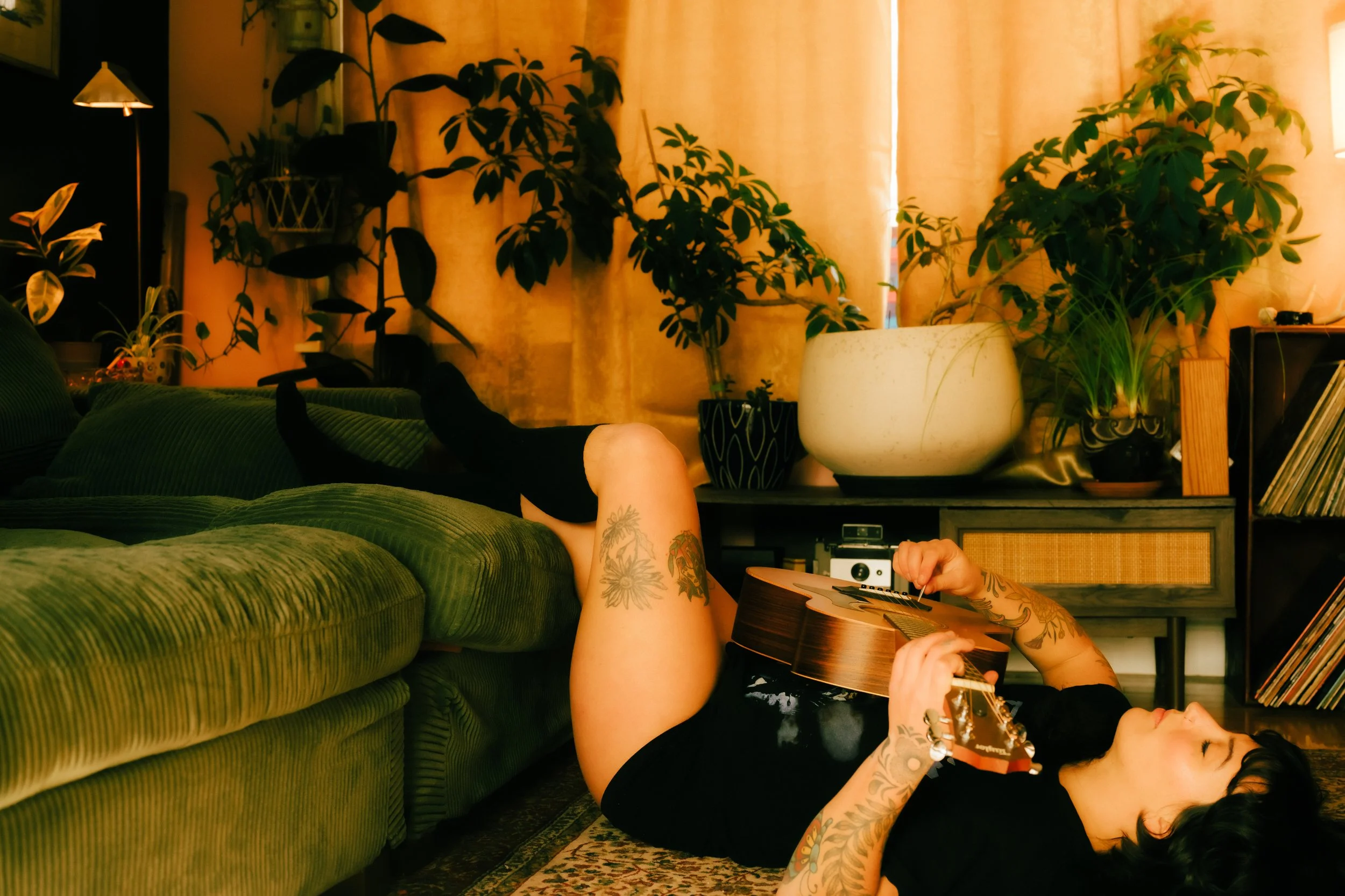 A woman with tattoos lies on her back on the floor playing an acoustic guitar, with her feet resting on a green couch in a cozy room decorated with potted plants and warm lighting.