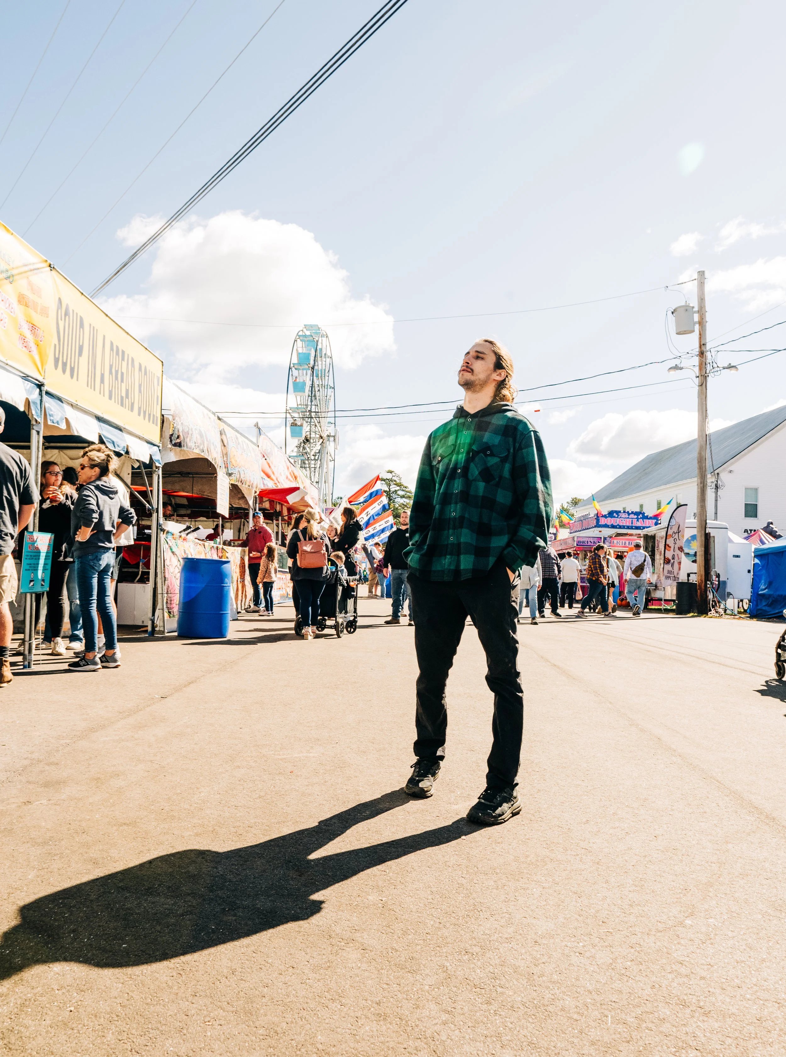 A young man in a green plaid shirt and black pants stands on a sunny day at a fairground with booths, a Ferris wheel, and other people in the background.