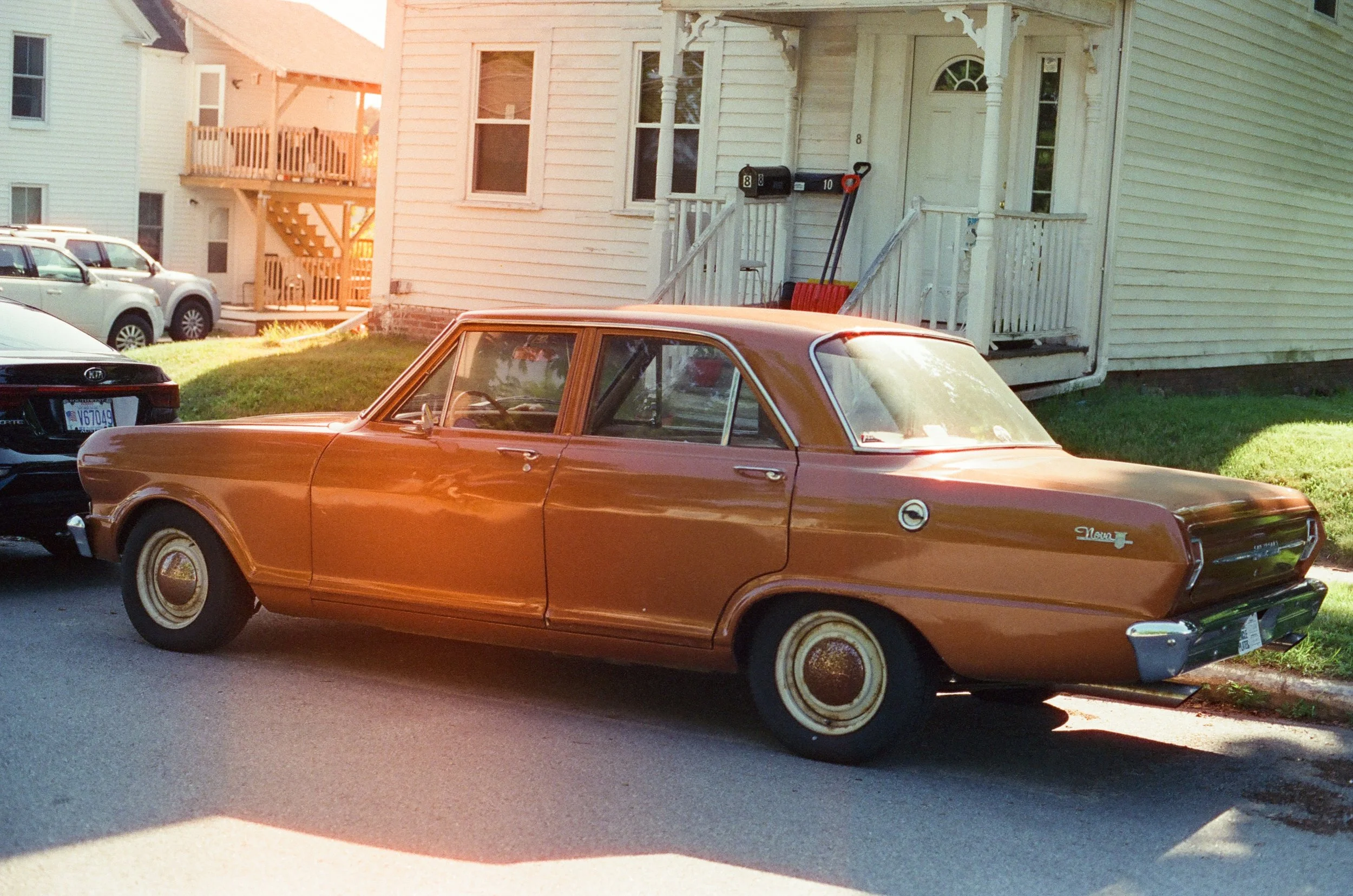 Vintage brown sedan parked on a residential street in front of a house with a small porch and mailbox.