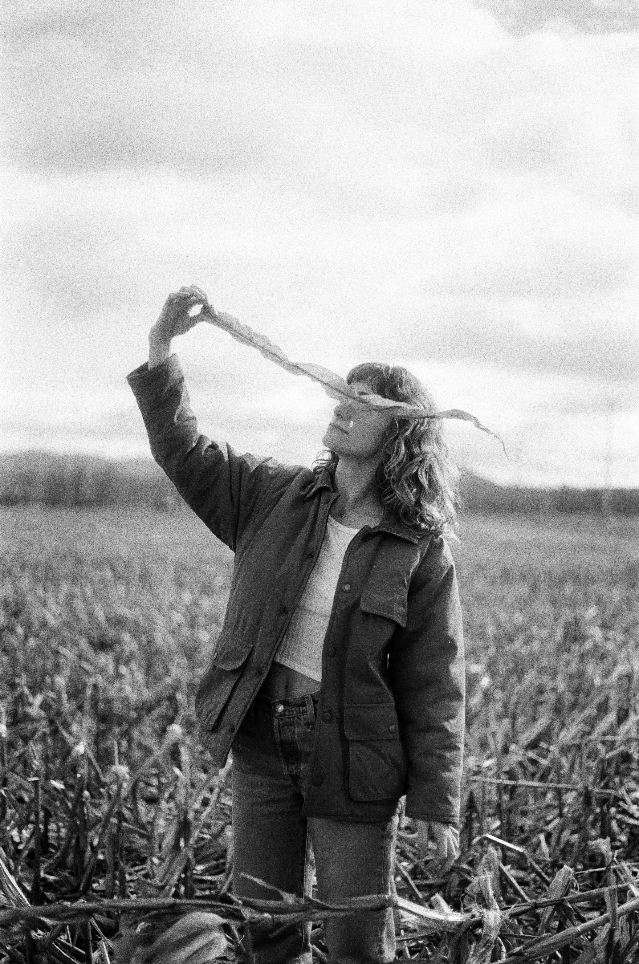 A woman with curly hair stands in a field of crops, holding a long piece of plant in front of her face, looking at it against an overcast sky.