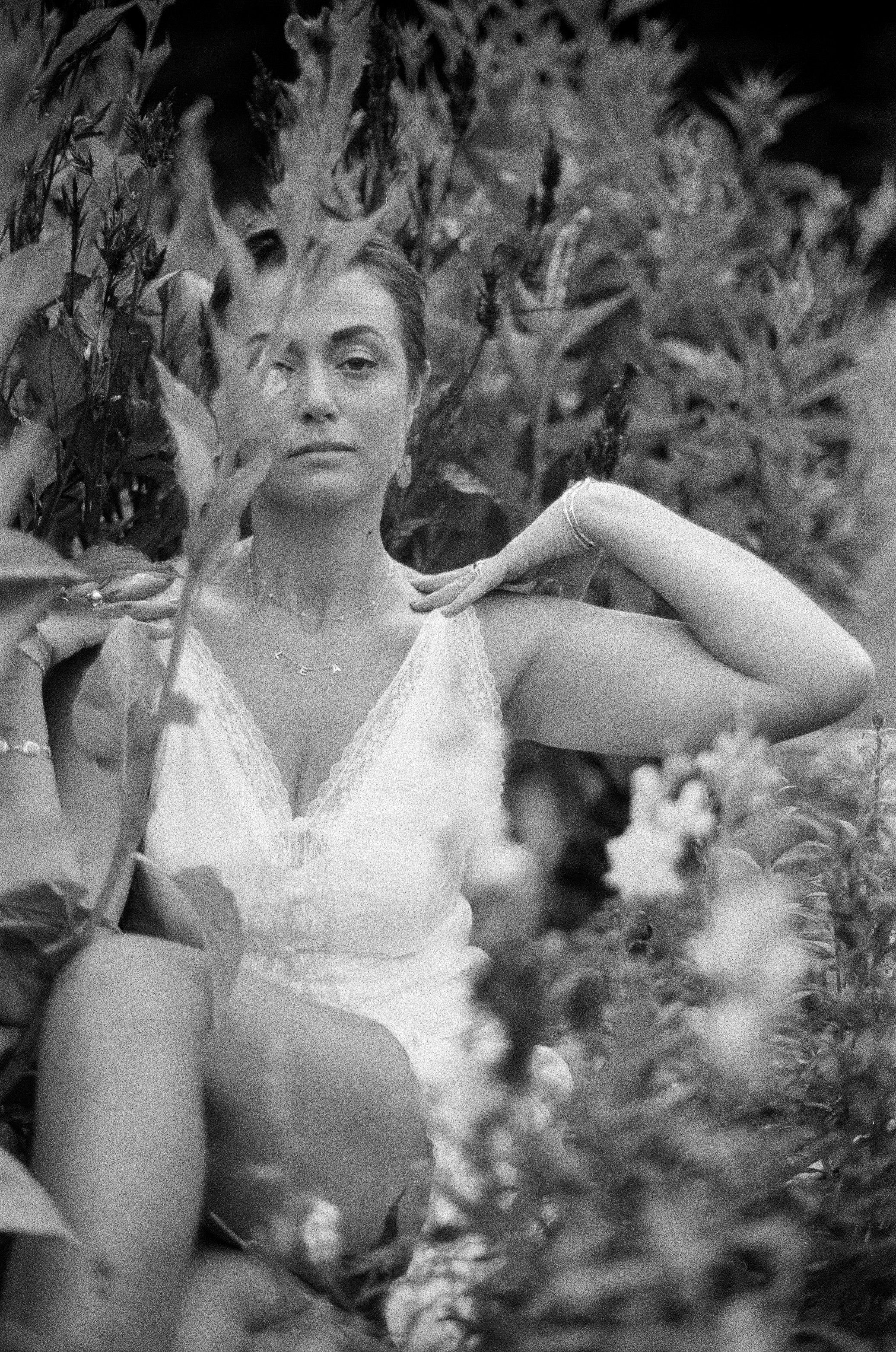 A woman in a white lace nightgown sitting among tall plants, looking at the camera with a serene expression.