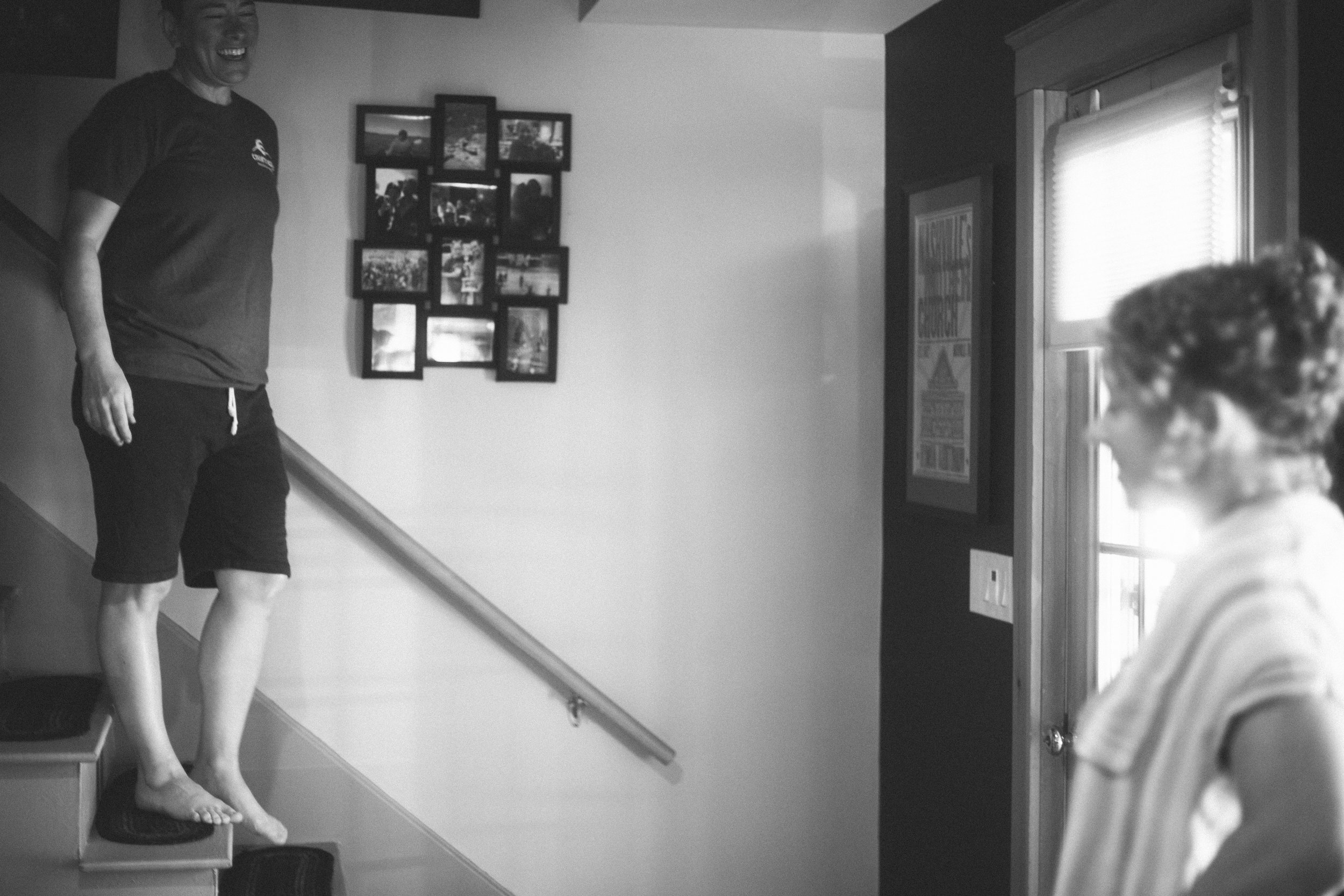 A woman is walking up a staircase with a smile, while another woman is seated near a window, looking toward her. There is a photo collage on the wall behind the woman on the stairs.