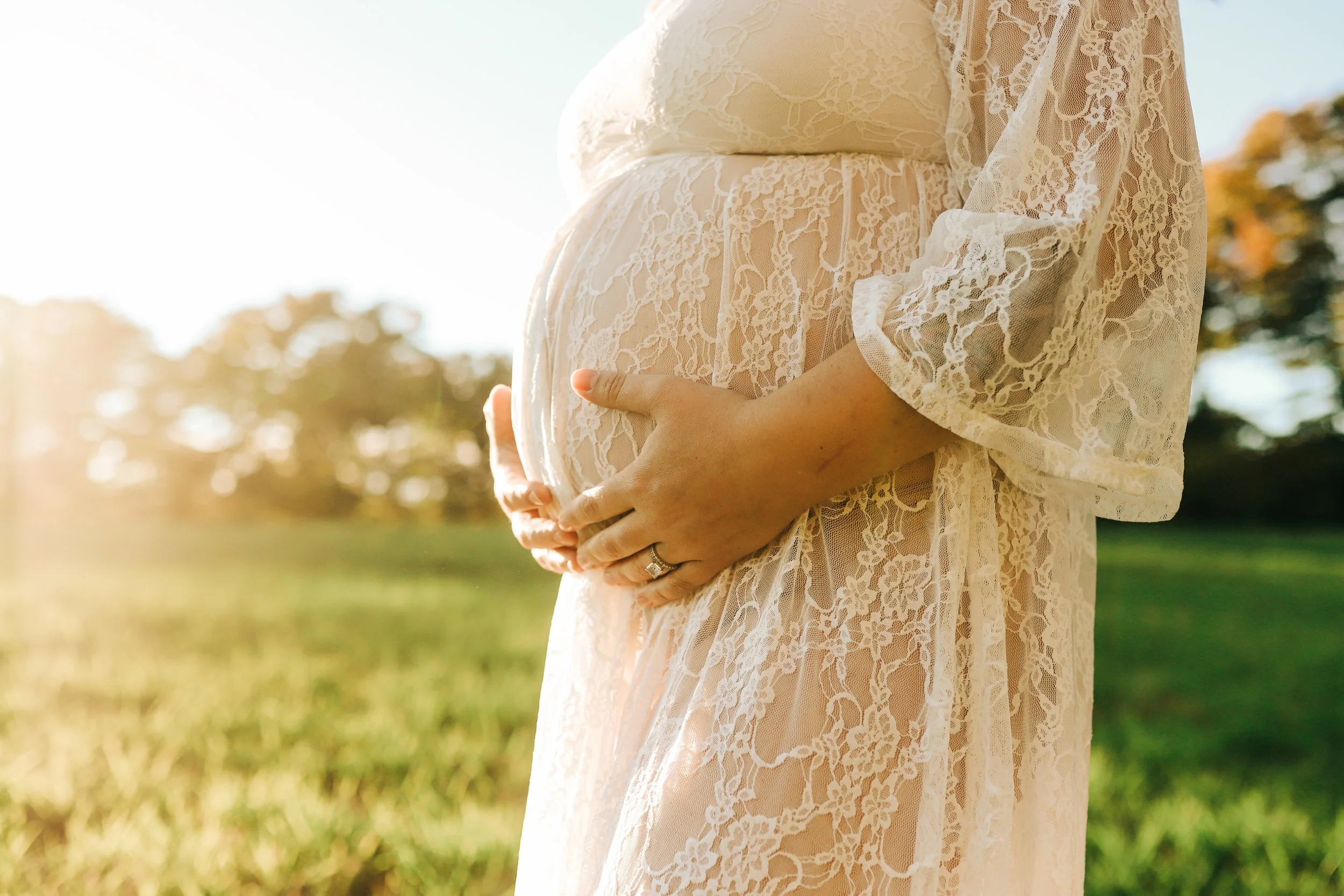 Close-up of a pregnant woman in a lace dress holding her belly with both hands, standing outdoors during sunlight with trees in the background.