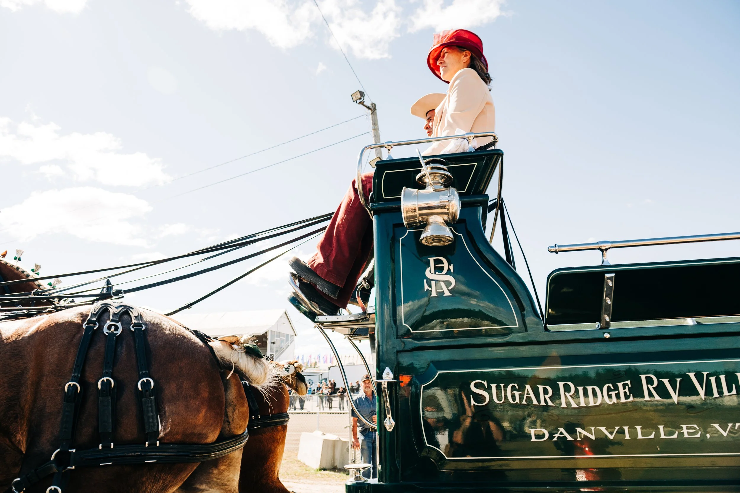 Women riding in a horse-drawn carriage with the sign "Sugar Ridge RV Village Danville VA." The carriage is dark green with chrome accents and a lantern, and the women wear hats, one in red and the other in beige. The sky is mostly clear with some clo
