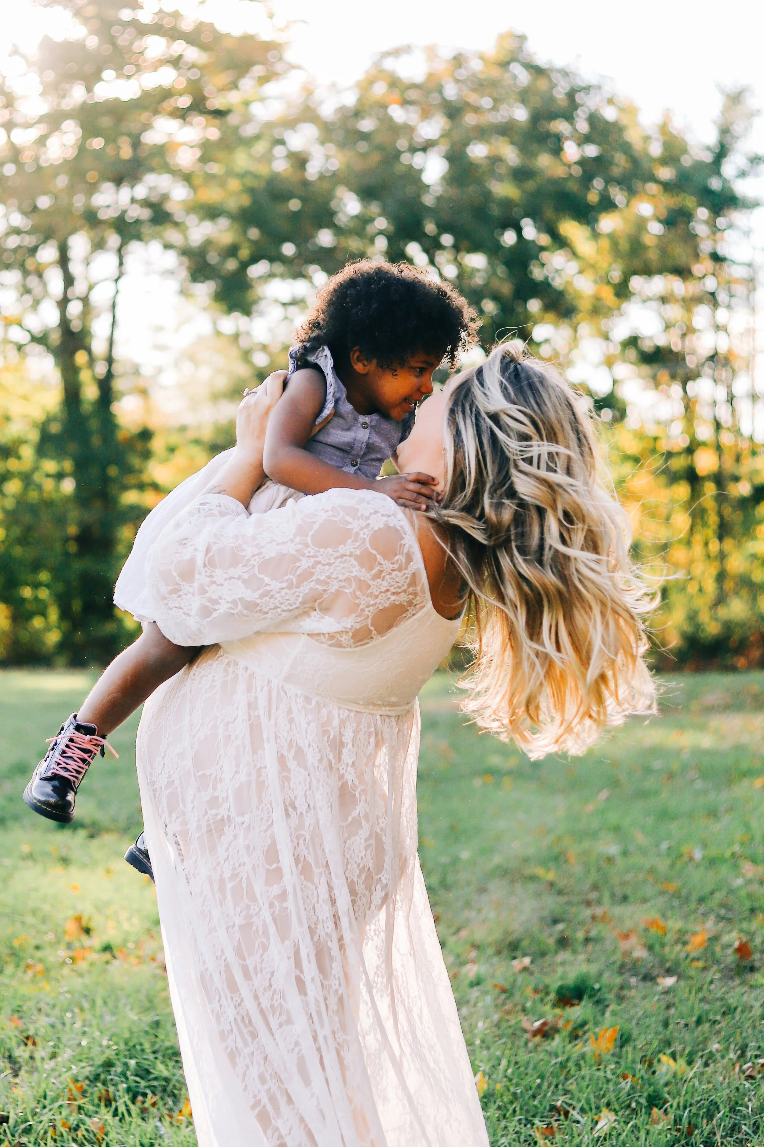 A woman in a lace dress kissing a young girl on the cheek in a park with sunlight filtering through trees.