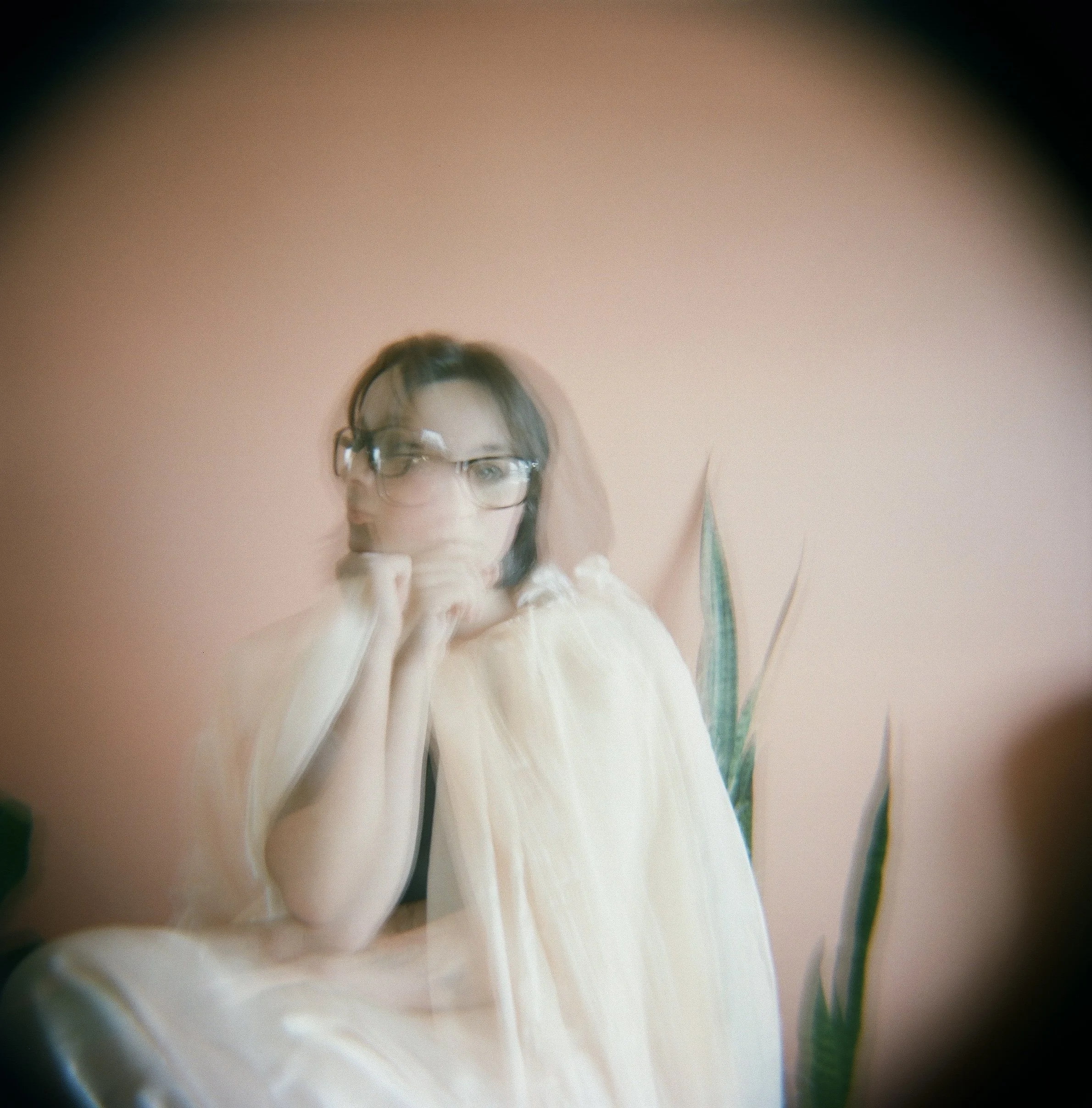 A girl with glasses and dark hair, resting her chin on her hand, seen through a circular lens or mirror with a soft pink background and a plant in the background.