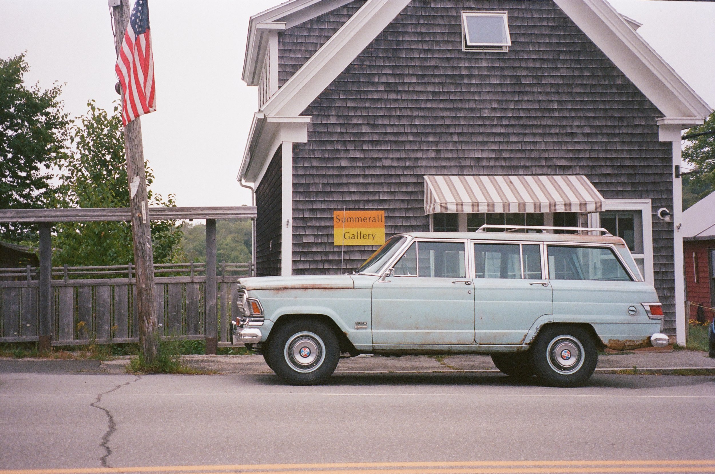 An old, light blue station wagon parked in front of a gray wooden building with a striped awning and a yellow sign that reads "Summerall Gallery". An American flag is on a pole attached to a wooden utility pole to the left of the car.