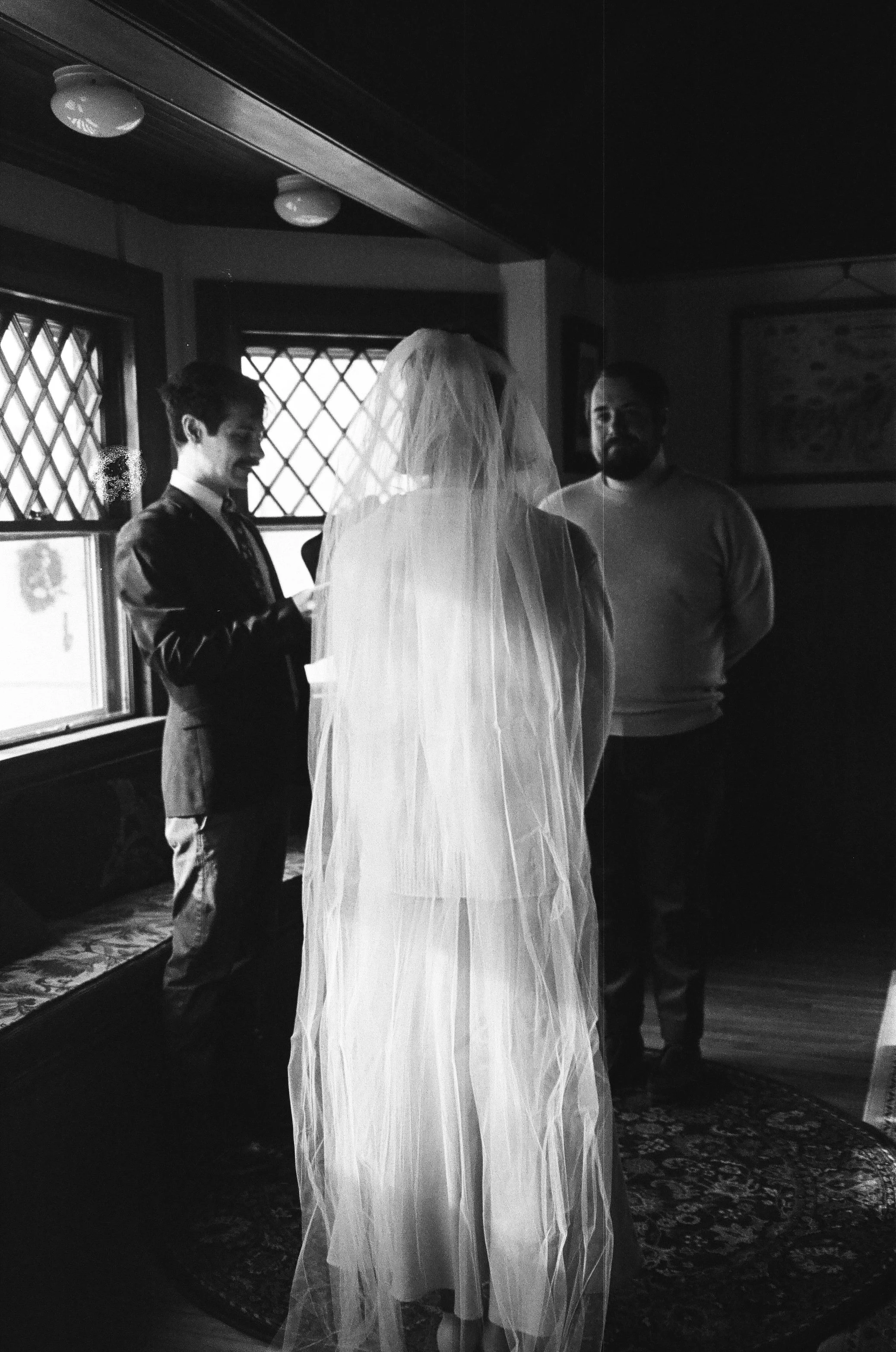 A woman dressed as a bride with a veil standing in front of a man in a suit and a man in a sweater inside a room with patterned windows and a rug.