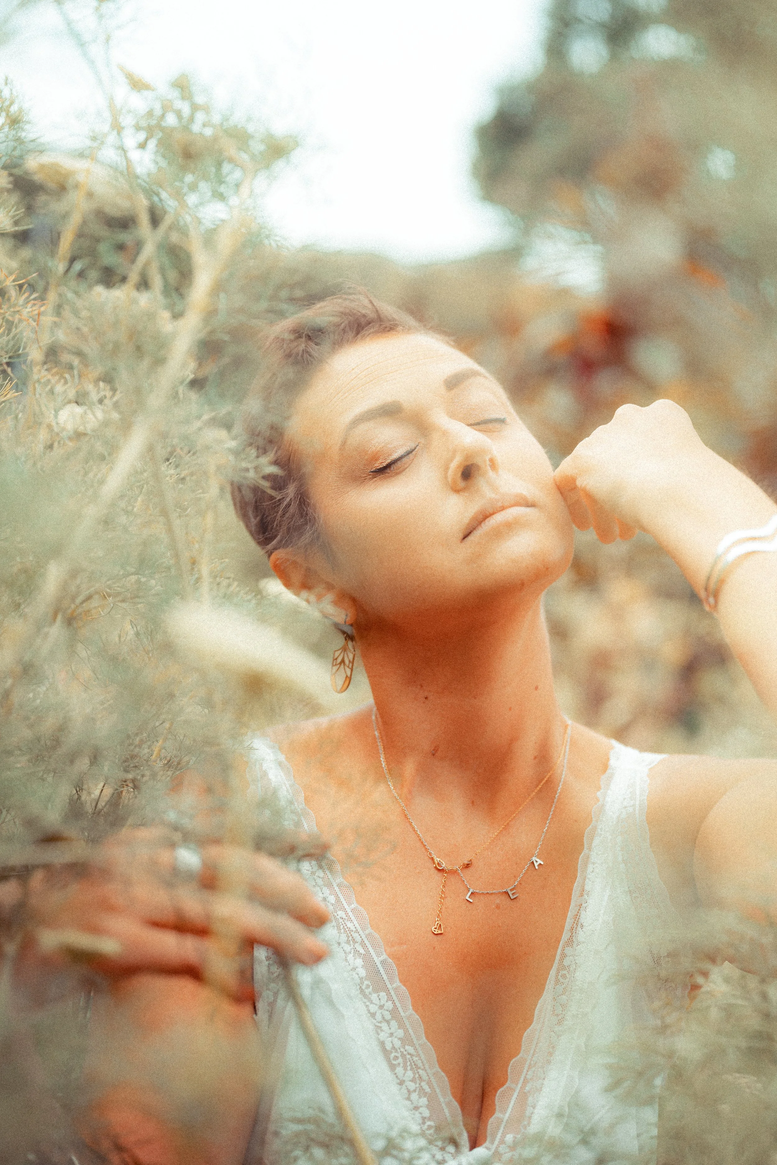 A woman with short hair and closed eyes, wearing a white lace top, gold necklace, earrings, and bracelets, is standing among plants with a calm expression.