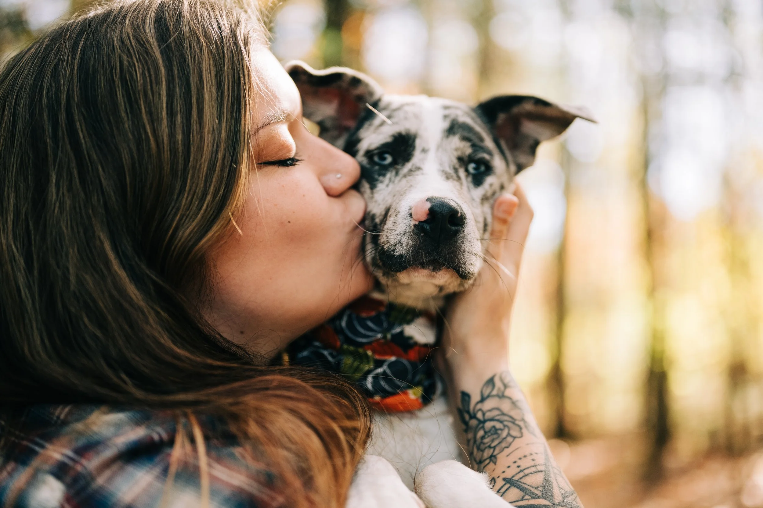 A woman with long brown hair is holding and kissing a blue merle Australian Shepherd puppy outdoors in a wooded area.