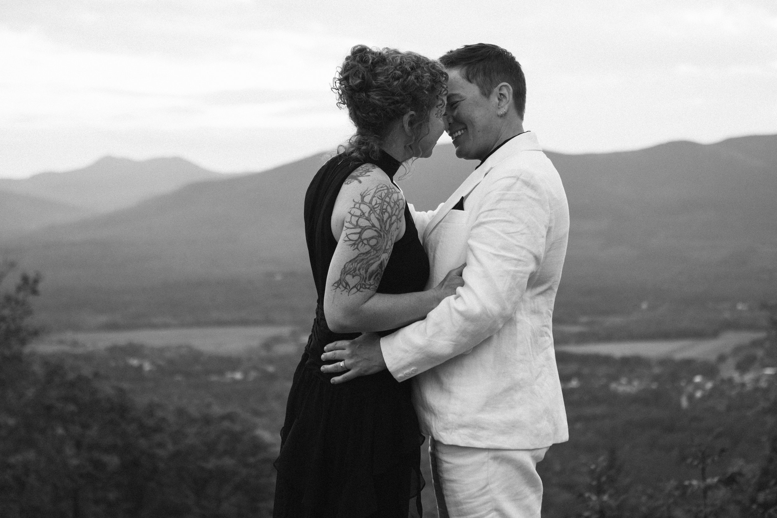A couple in wedding attire sharing a romantic moment outdoors with hills and mountains in the background.