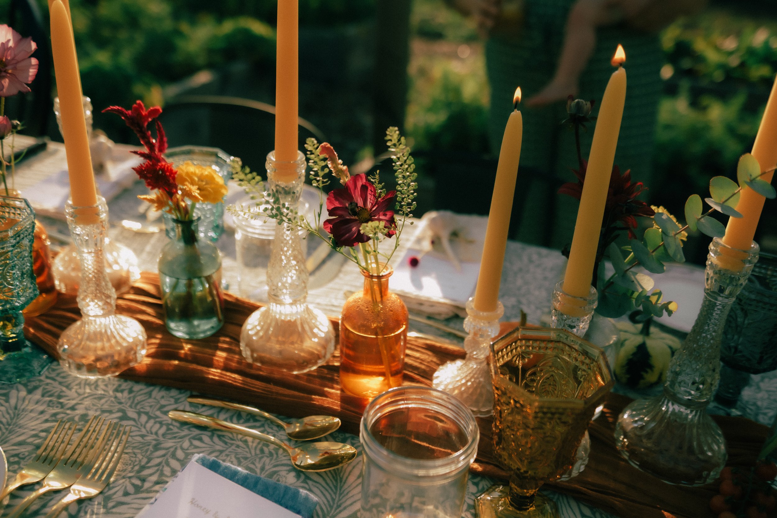 Table set for a celebration with candles, flowers in vases, and gold utensils, outdoors with greenery in the background.