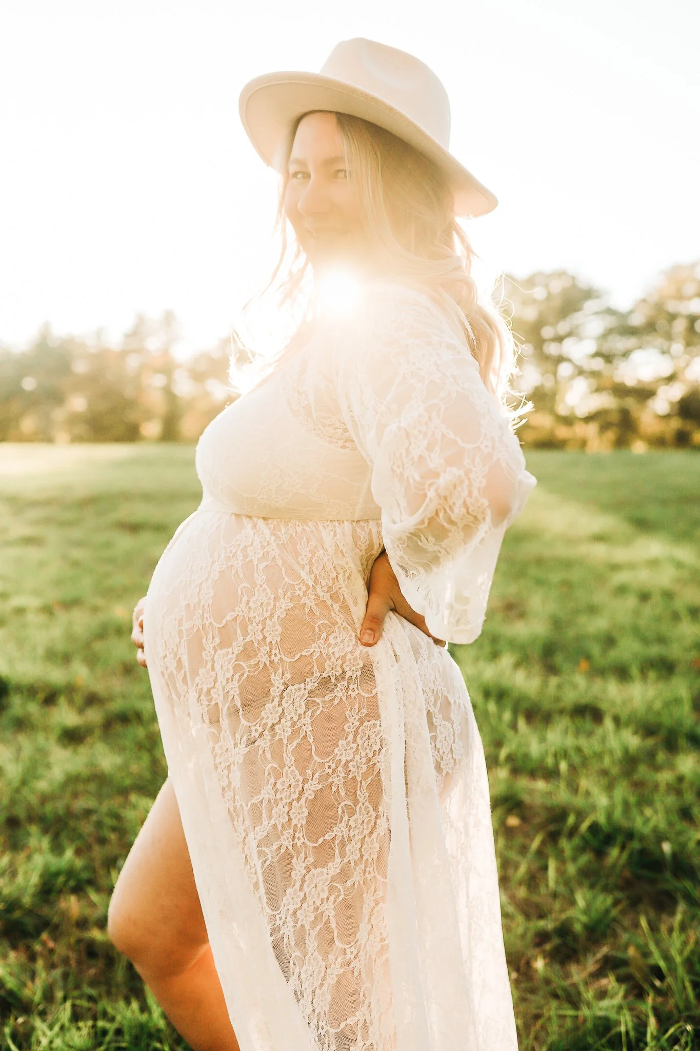 Pregnant woman smiling outdoors wearing a white lace dress and wide-brimmed hat during sunset.
