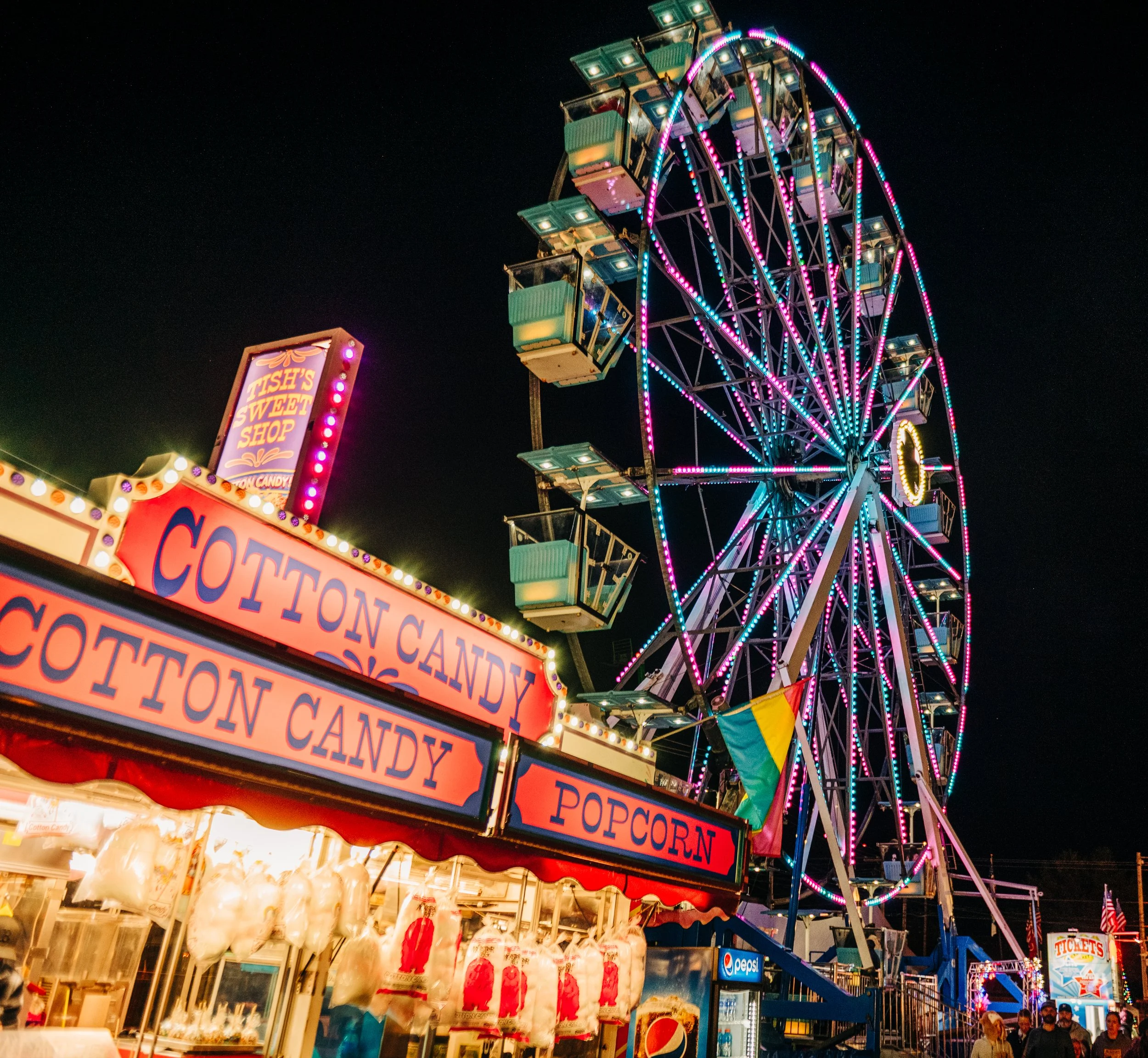 Nighttime scene at a carnival with a large illuminated Ferris wheel and booth selling cotton candy, popcorn, and candy, with colorful lights and people walking.