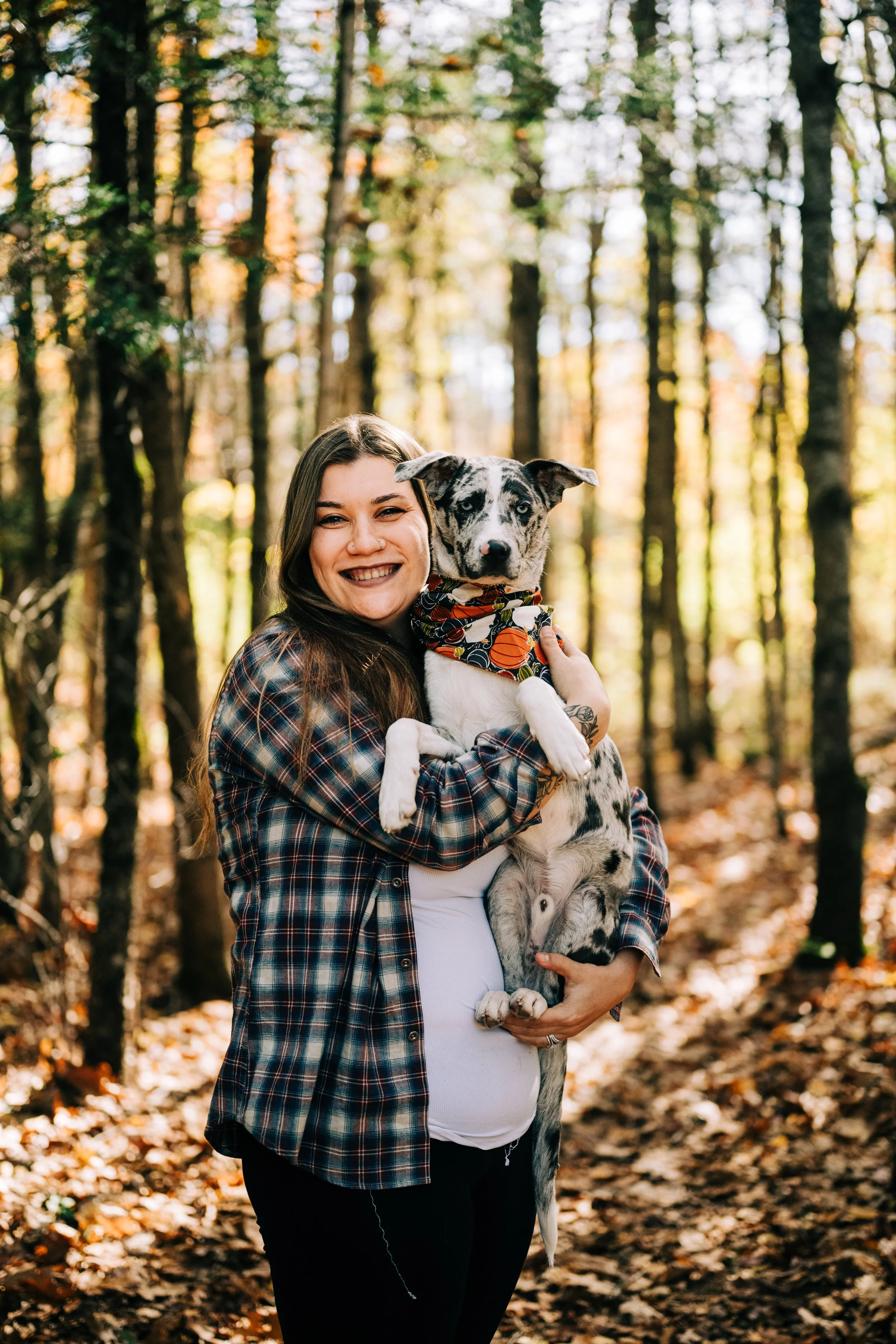 A woman in a plaid shirt holding a black and white mixed breed dog in a forest with autumn leaves.