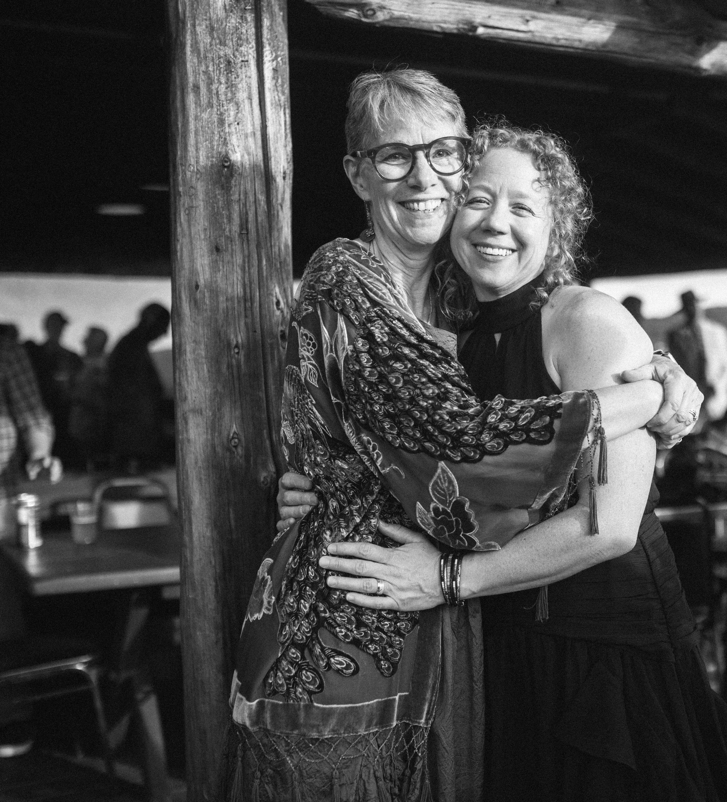 Two women hugging and smiling in a social setting, with a rustic wooden pillar behind them and blurred silhouettes of people in the background.