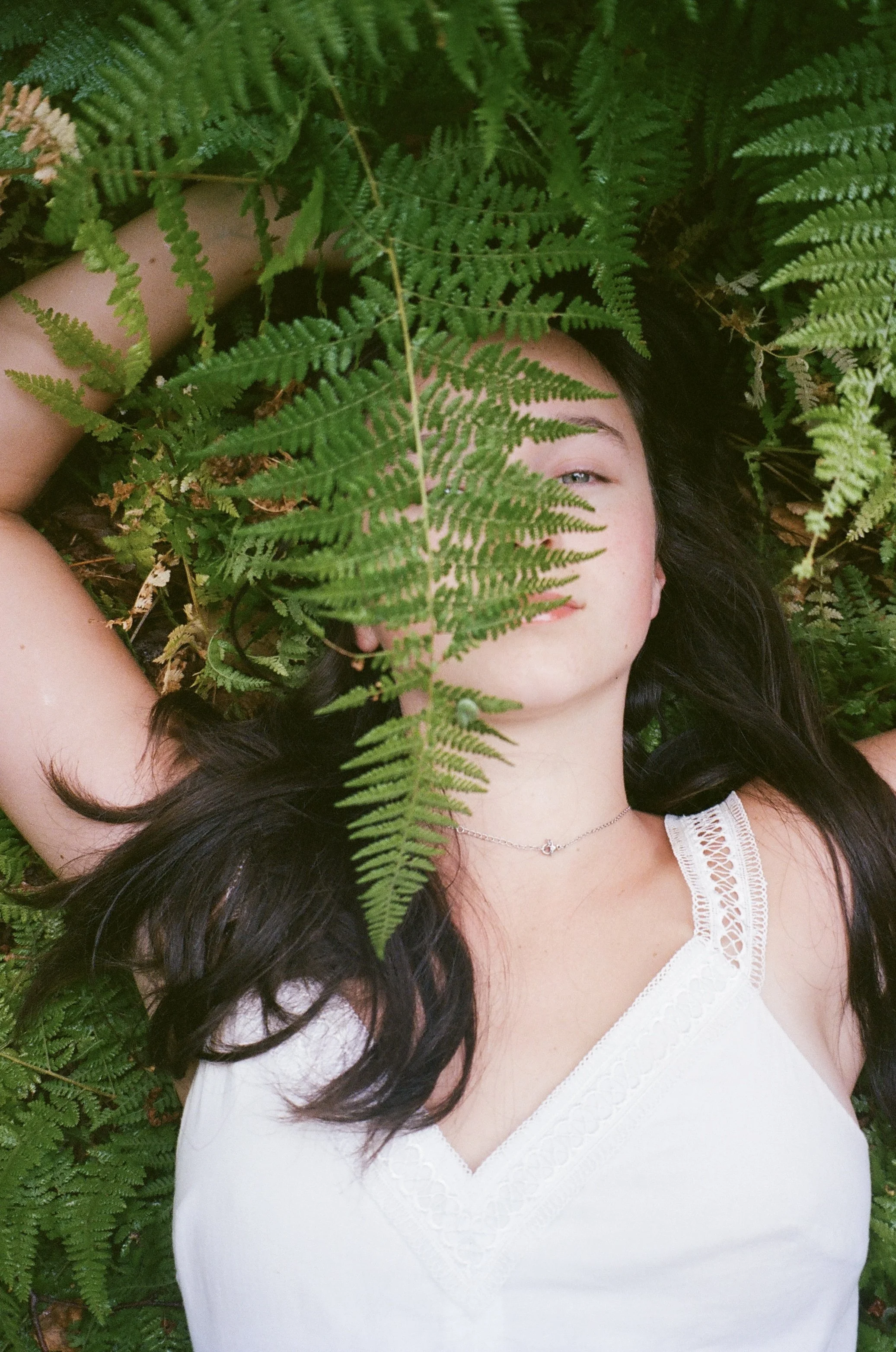 A woman lying among green fern leaves, partially covering her face and body, wearing a white lace-trimmed top and a delicate necklace.
