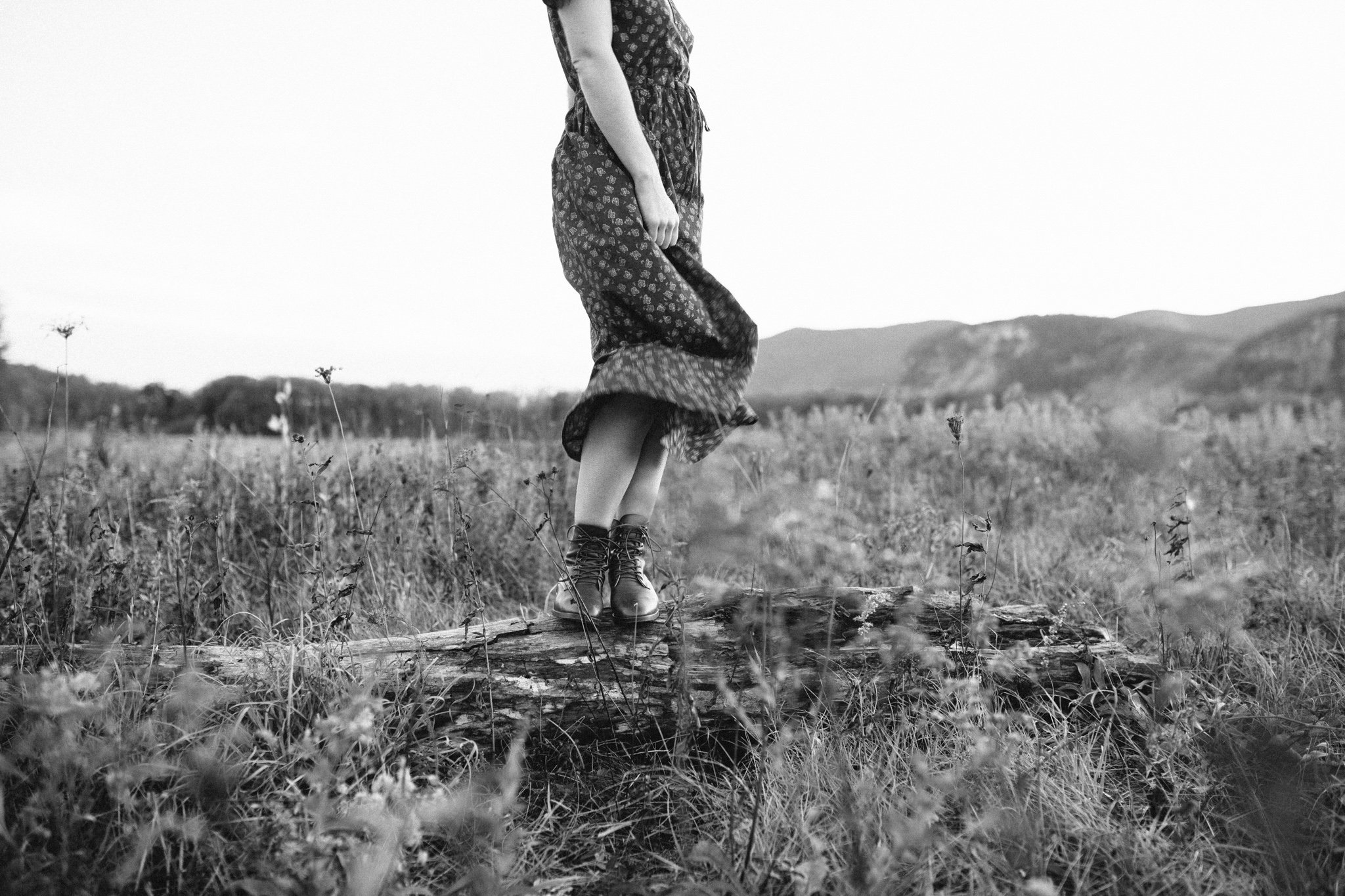 Black and white photo of a person standing on a tree trunk in a field, wearing a dress and boots, with mountains in the background.