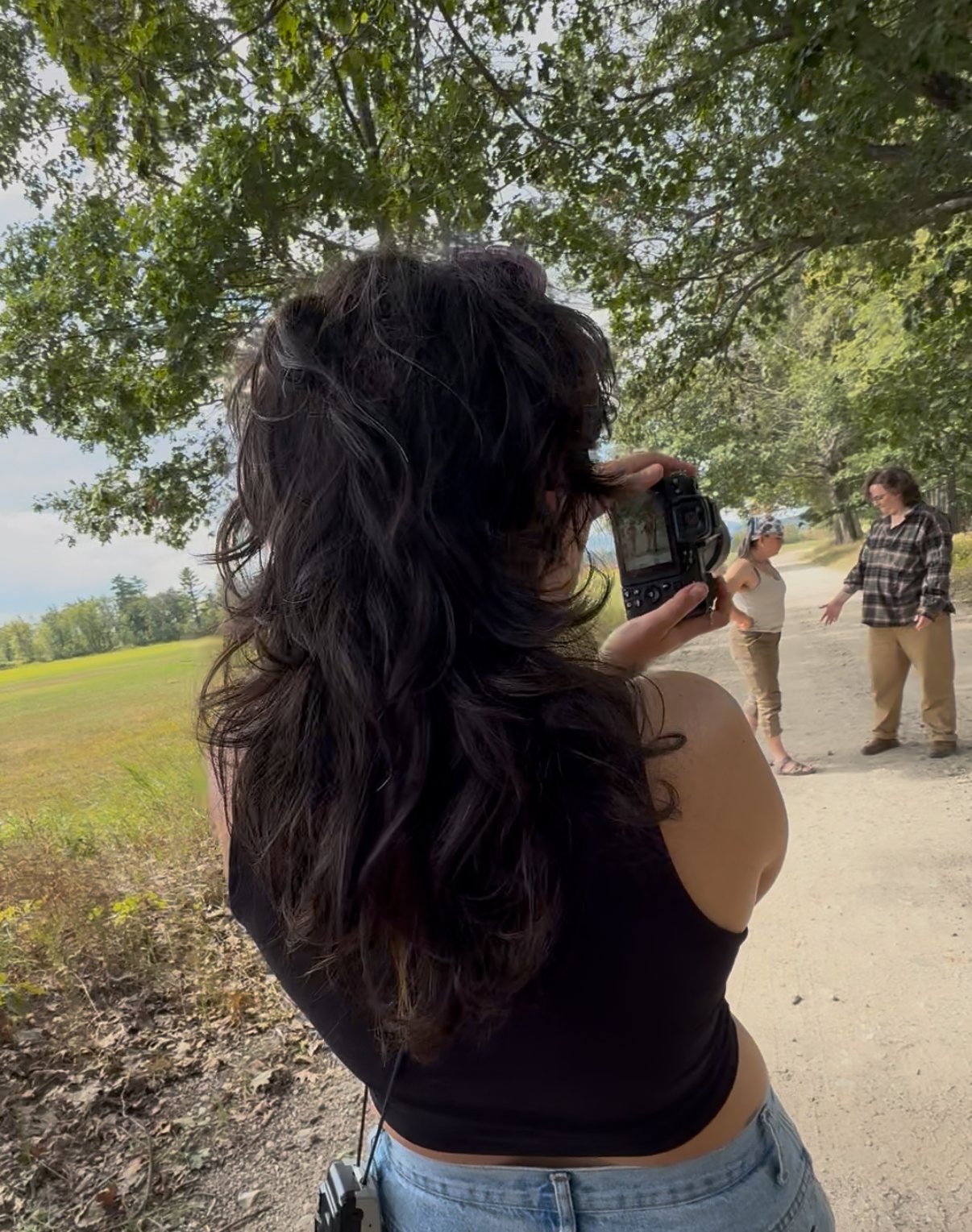 A woman with long, curly dark hair takes a photo with a camera of two other women who are outdoors under a large tree.